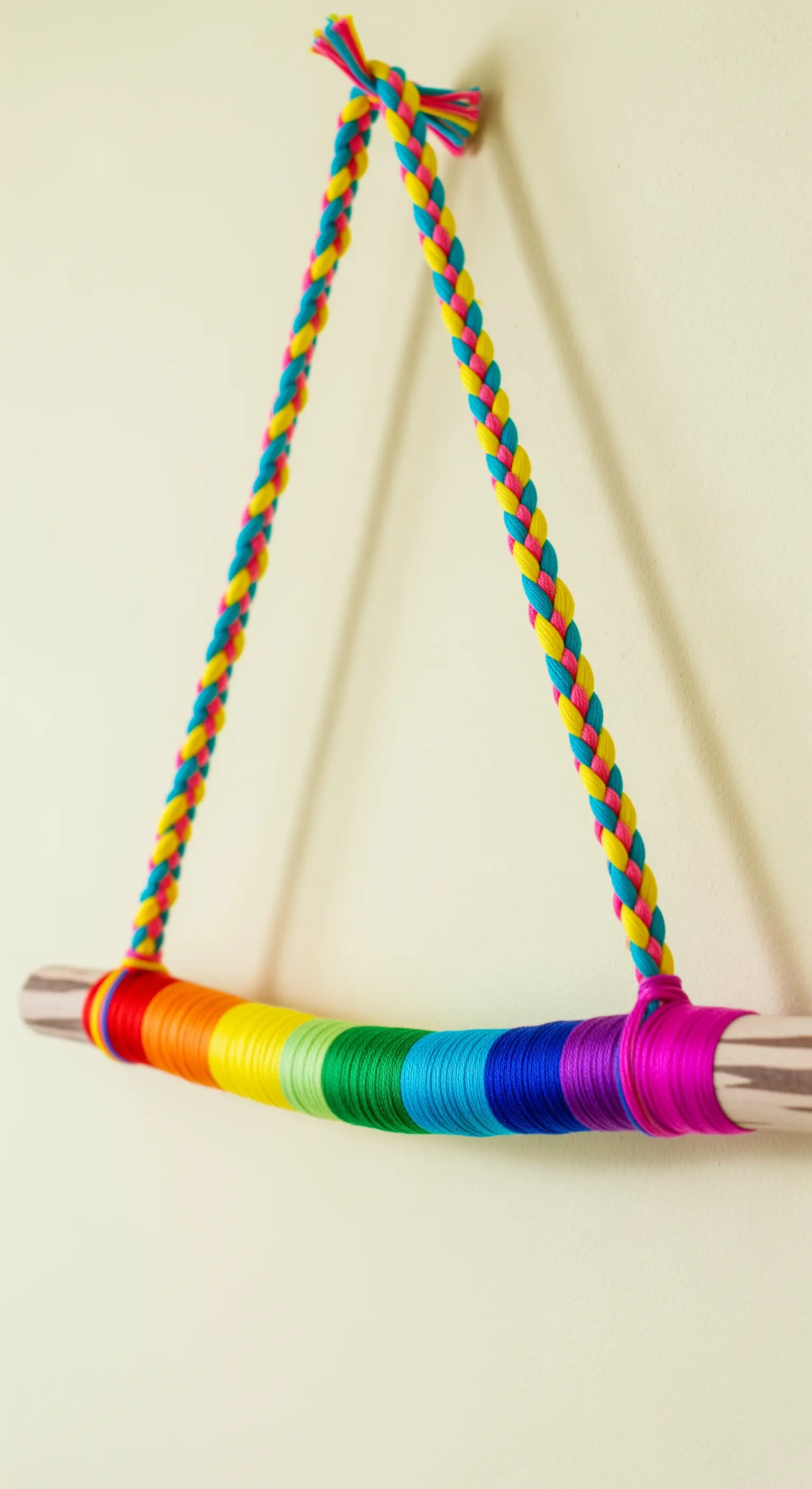Bastone di legno avvolto in fili di cotone color arcobaleno, appeso con una corda intrecciata.