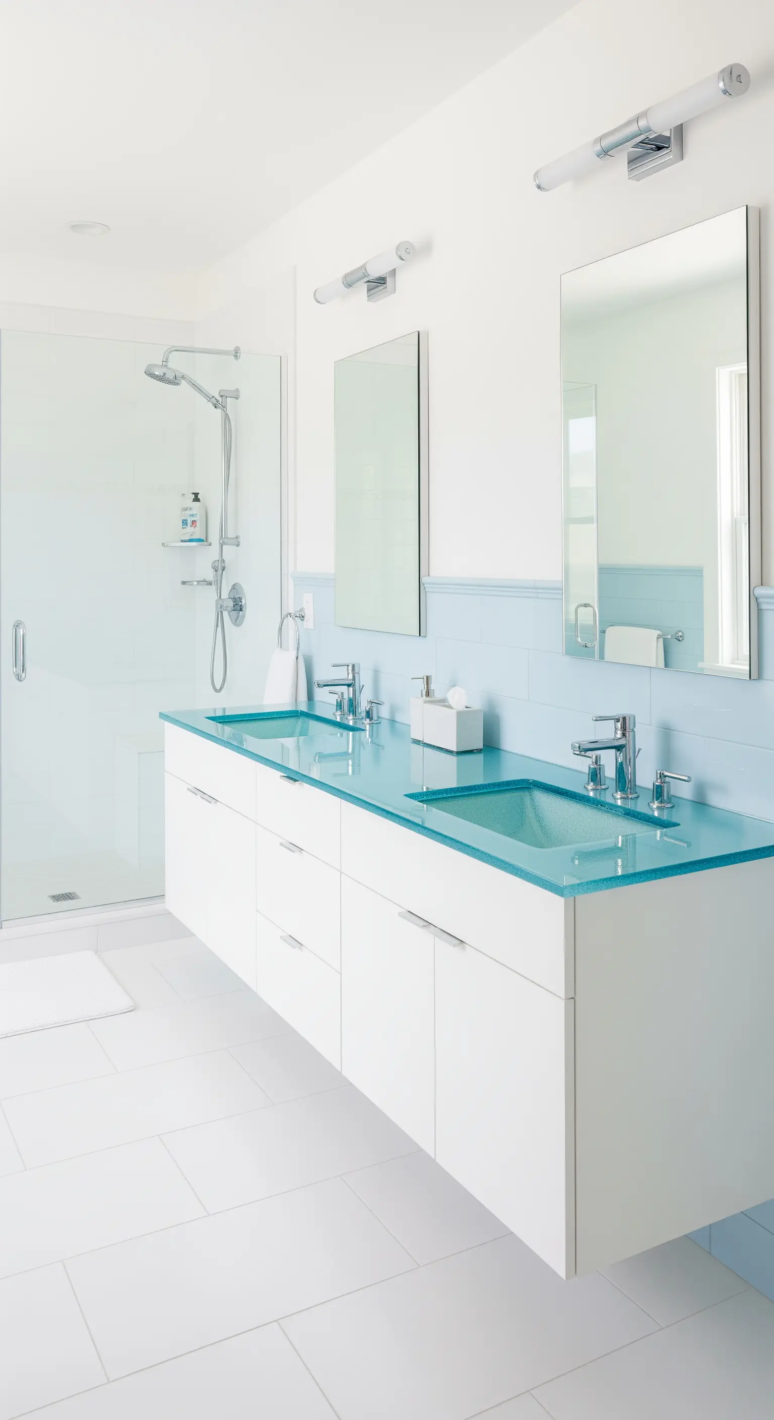 Bagno luminoso con mezza parete piastrellata di azzurro e top in vetro coordinato.
