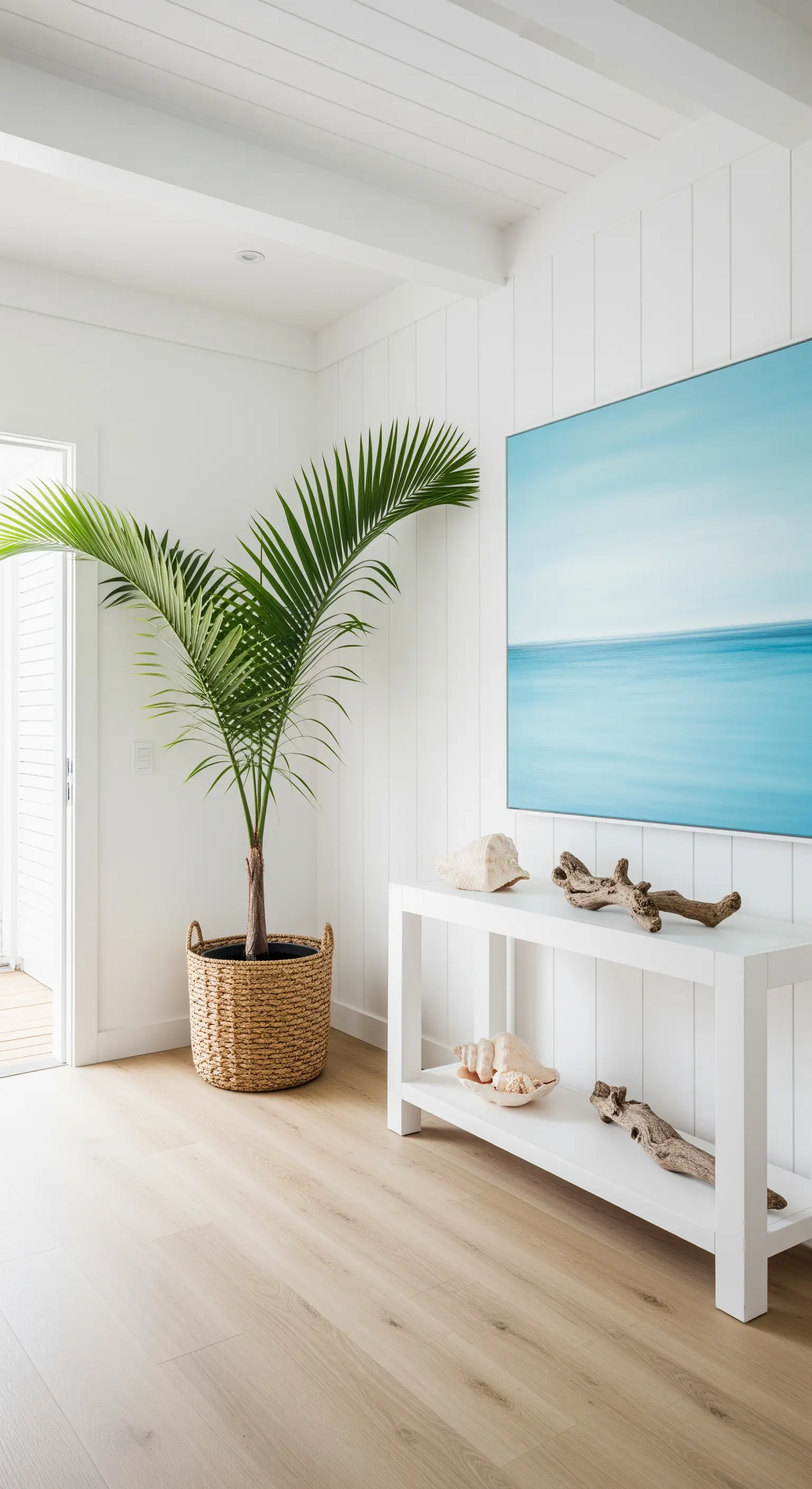 Palma Kentia in un cesto di vimini in un ingresso bianco in stile costiero con pavimento in legno chiaro.