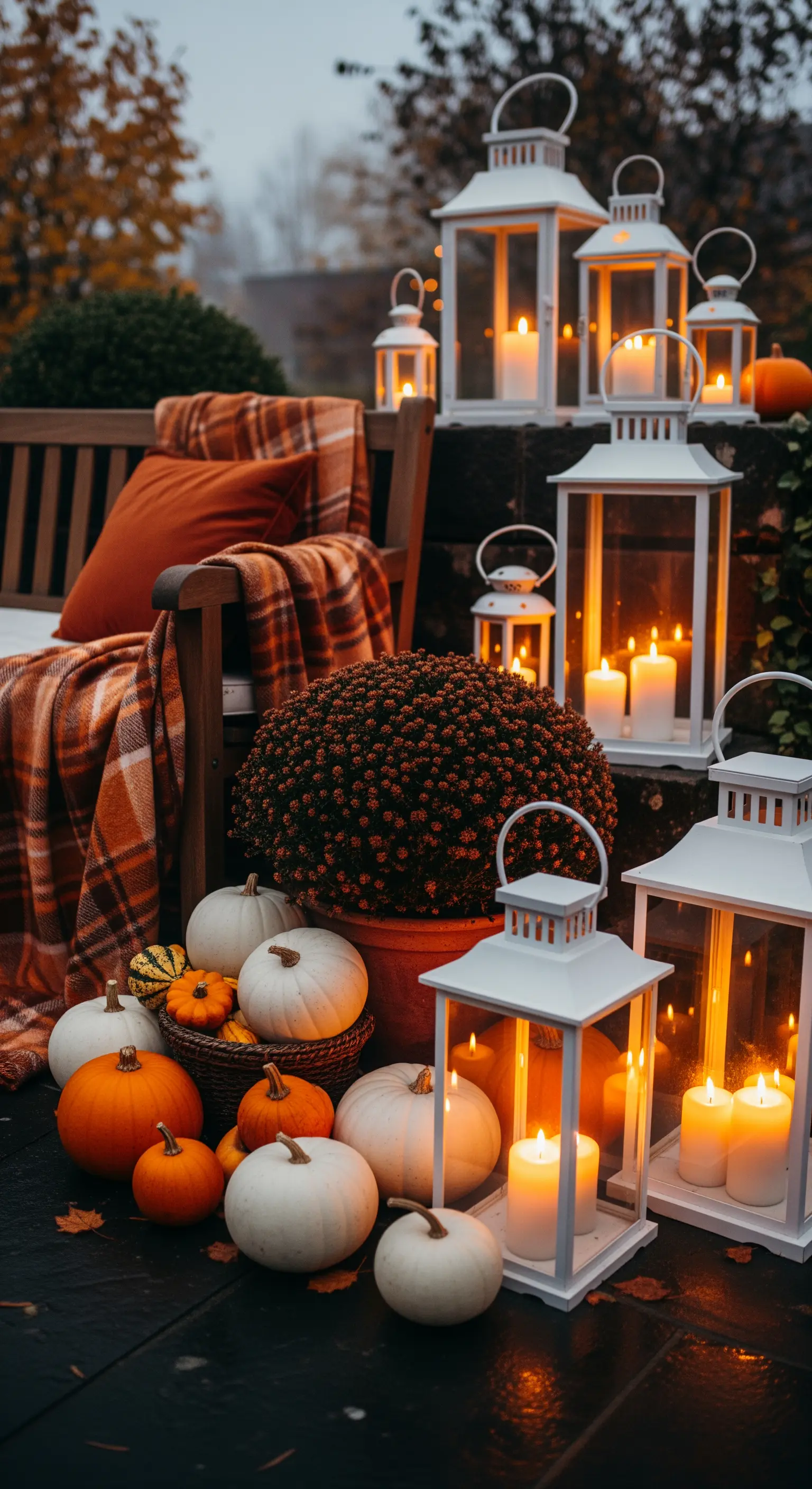 Composizione autunnale con lanterne bianche accese, zucche, fiori e plaid arancione.