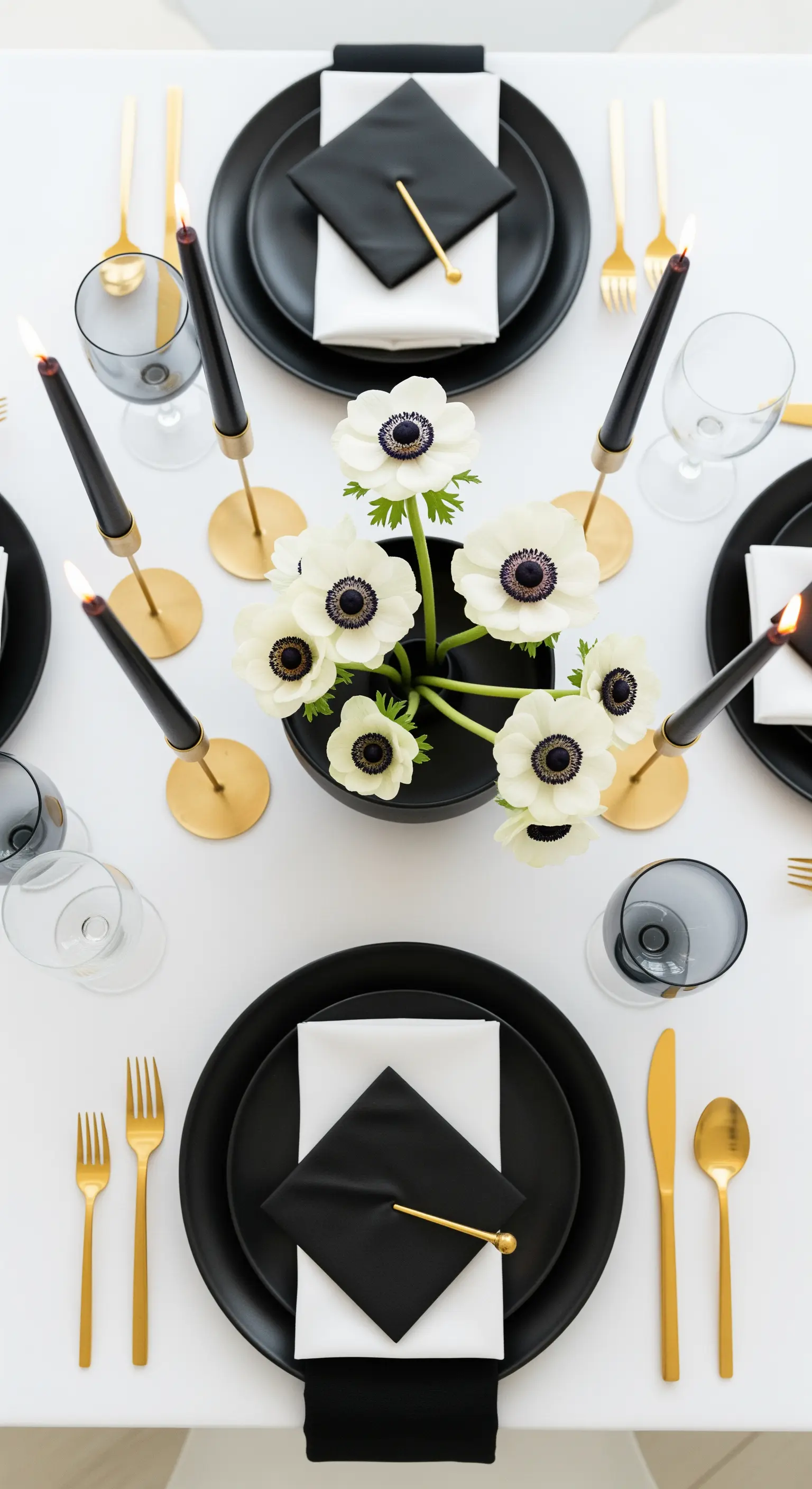 Tavola minimalista con piatti neri, posate dorate, candele nere e anemoni bianchi.