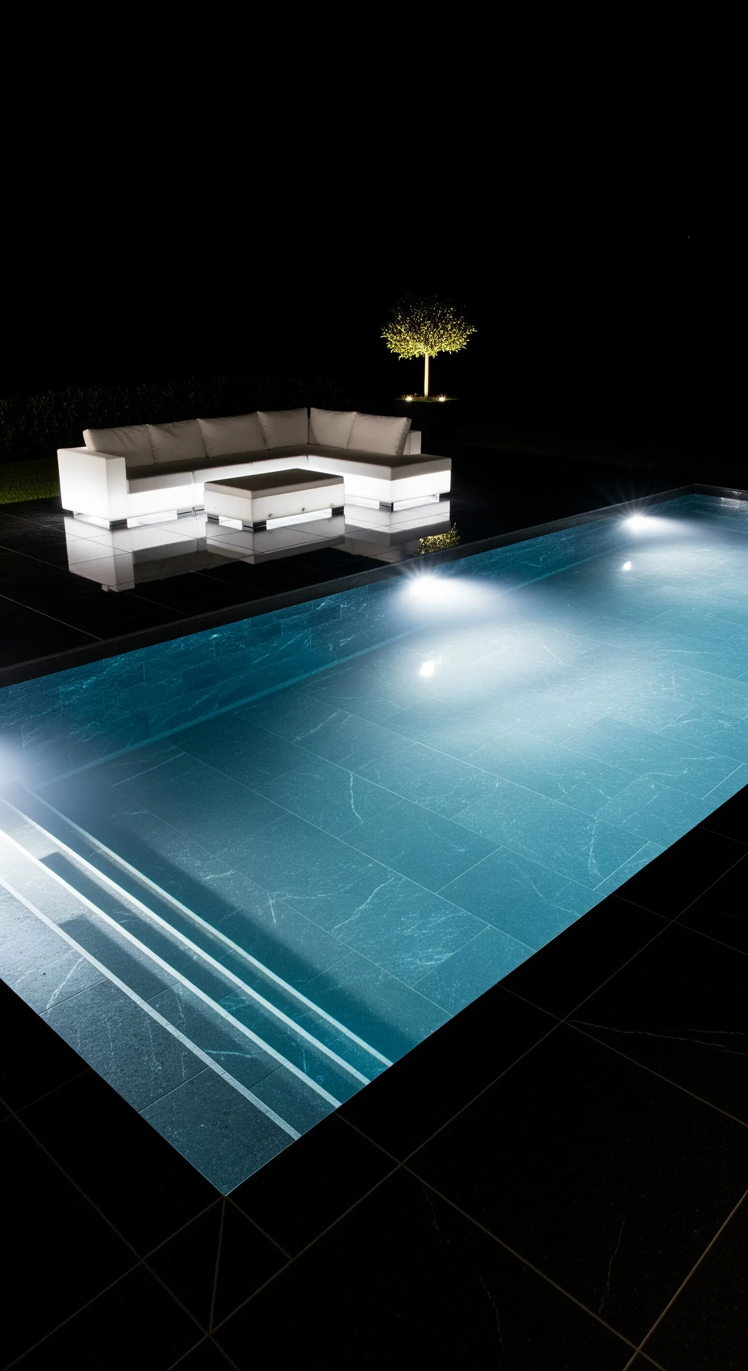 Area piscina minimalista e scura di notte con un divano e un tavolo bianchi che si illuminano dall'interno.