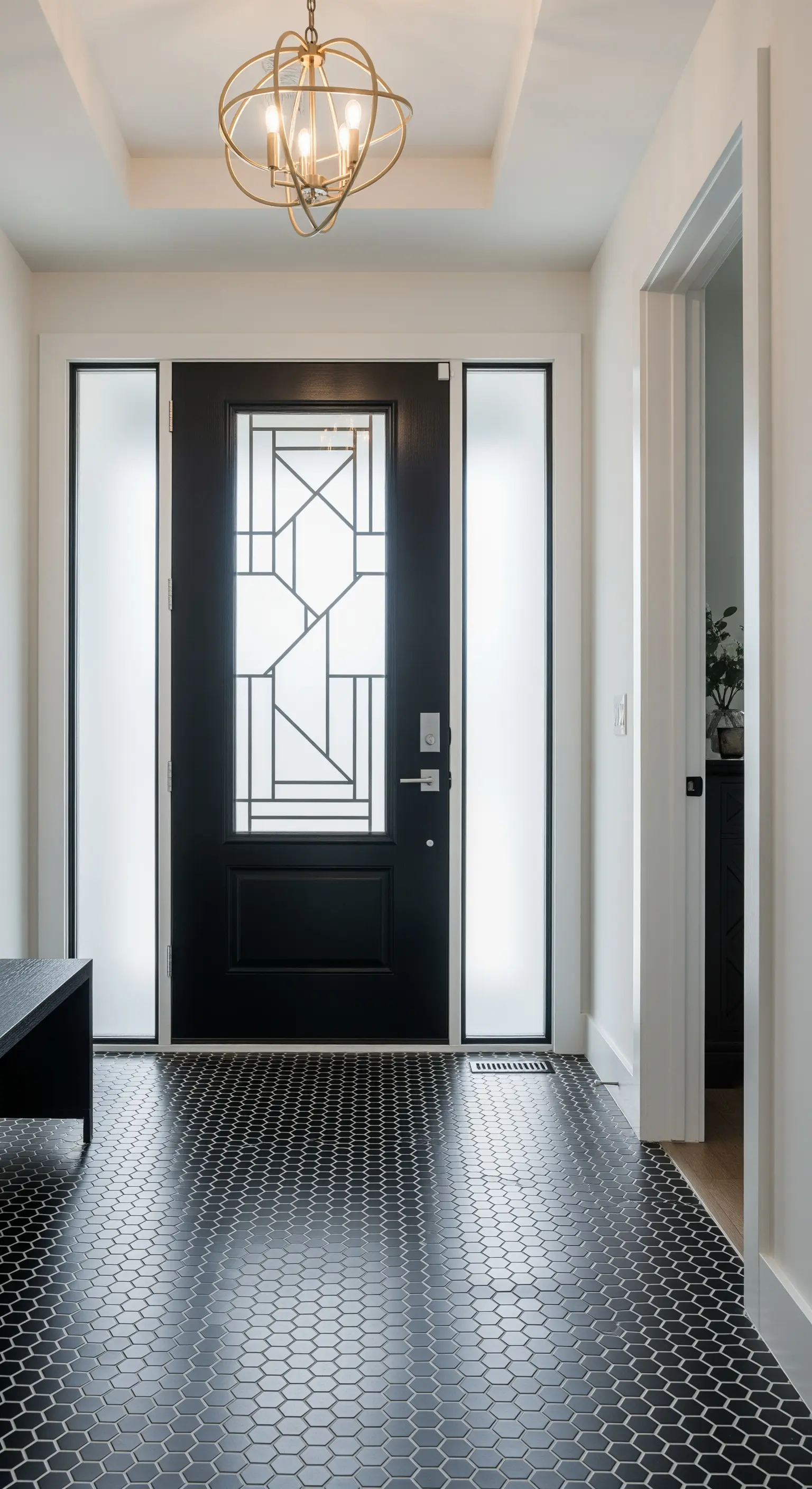 Porta d'ingresso nera con vetri geometrici e pavimento in piastrelle esagonali nere con fughe bianche.