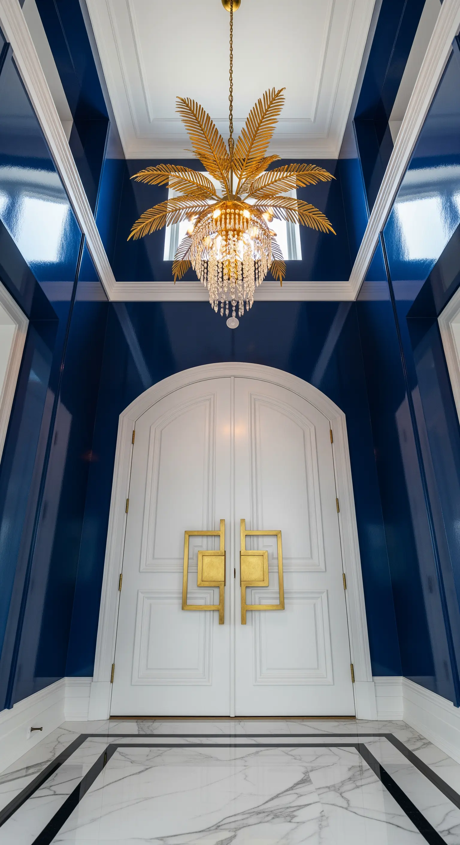 Ingresso stretto e alto con pareti laccate blu, porta bianca e lampadario dorato a forma di palma.