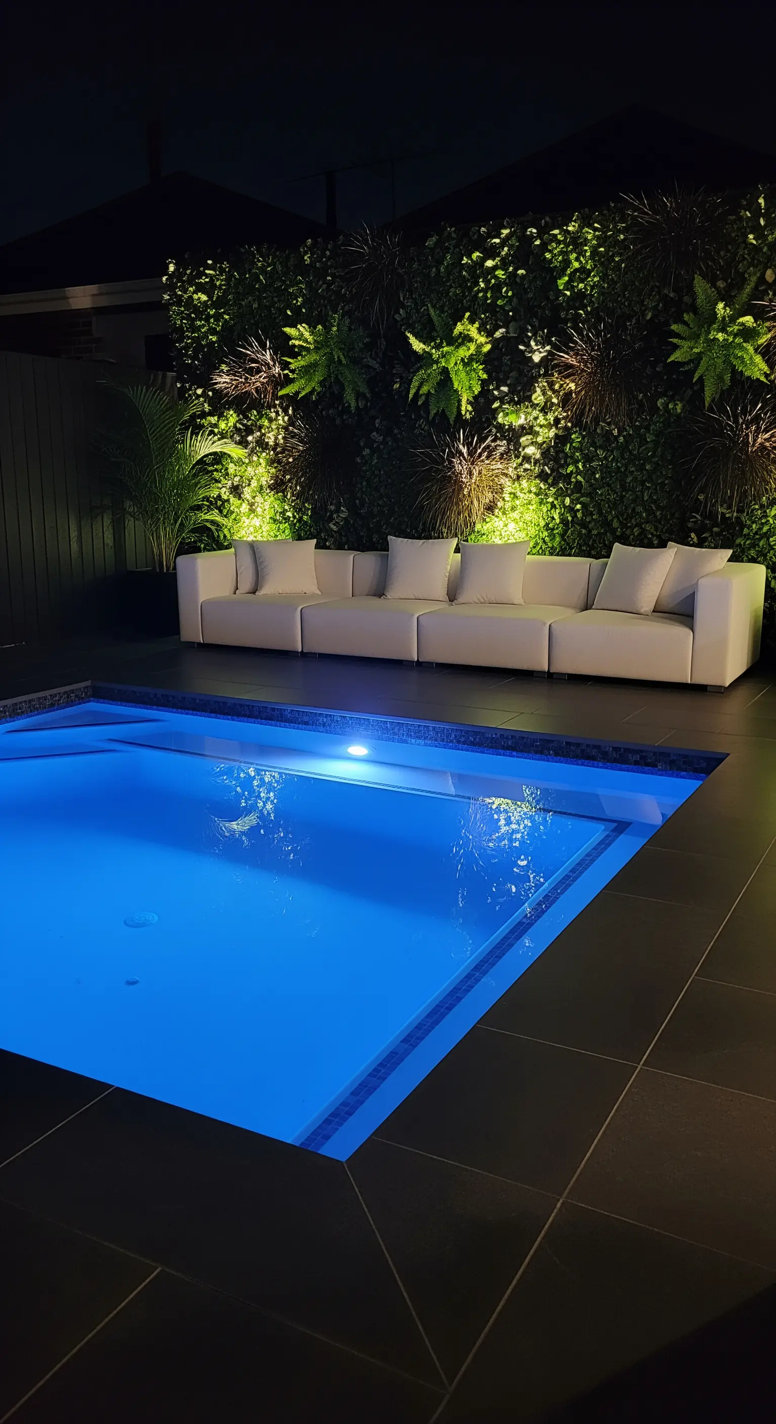 Piscina illuminata di notte con divano in pelle bianca e parete verde con faretti.