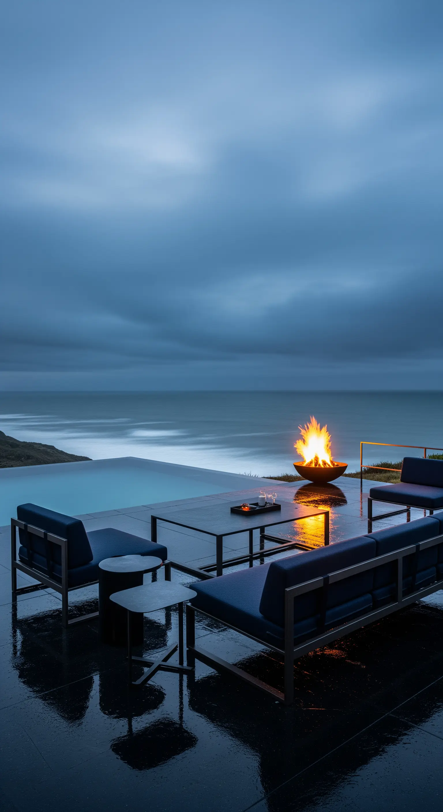 Salotto outdoor minimalista scuro con braciere acceso vicino a una piscina a sfioro sull'oceano.