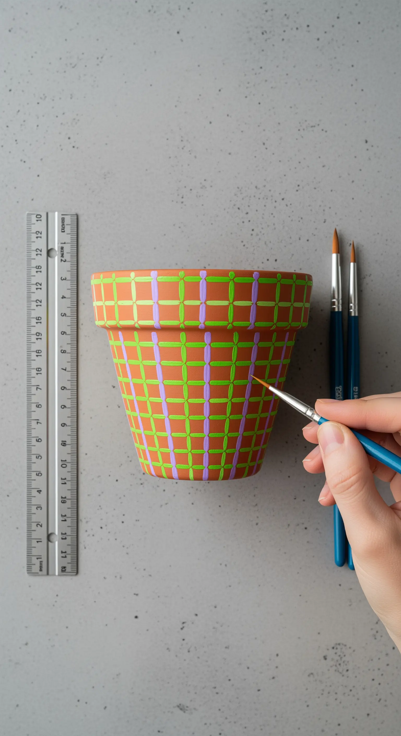 Mano che dipinge una griglia di linee verdi e lilla su un vaso di terracotta, creando un effetto tessuto.