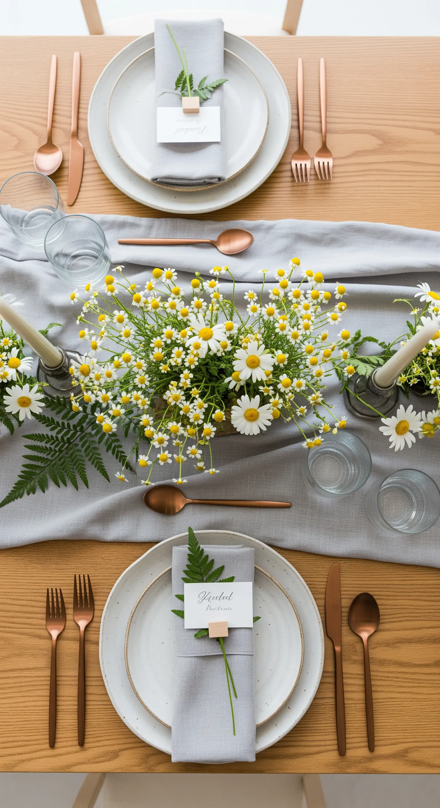 Vista dall'alto di una tavola con runner grigio, mazzi di camomilla e posate in rame.