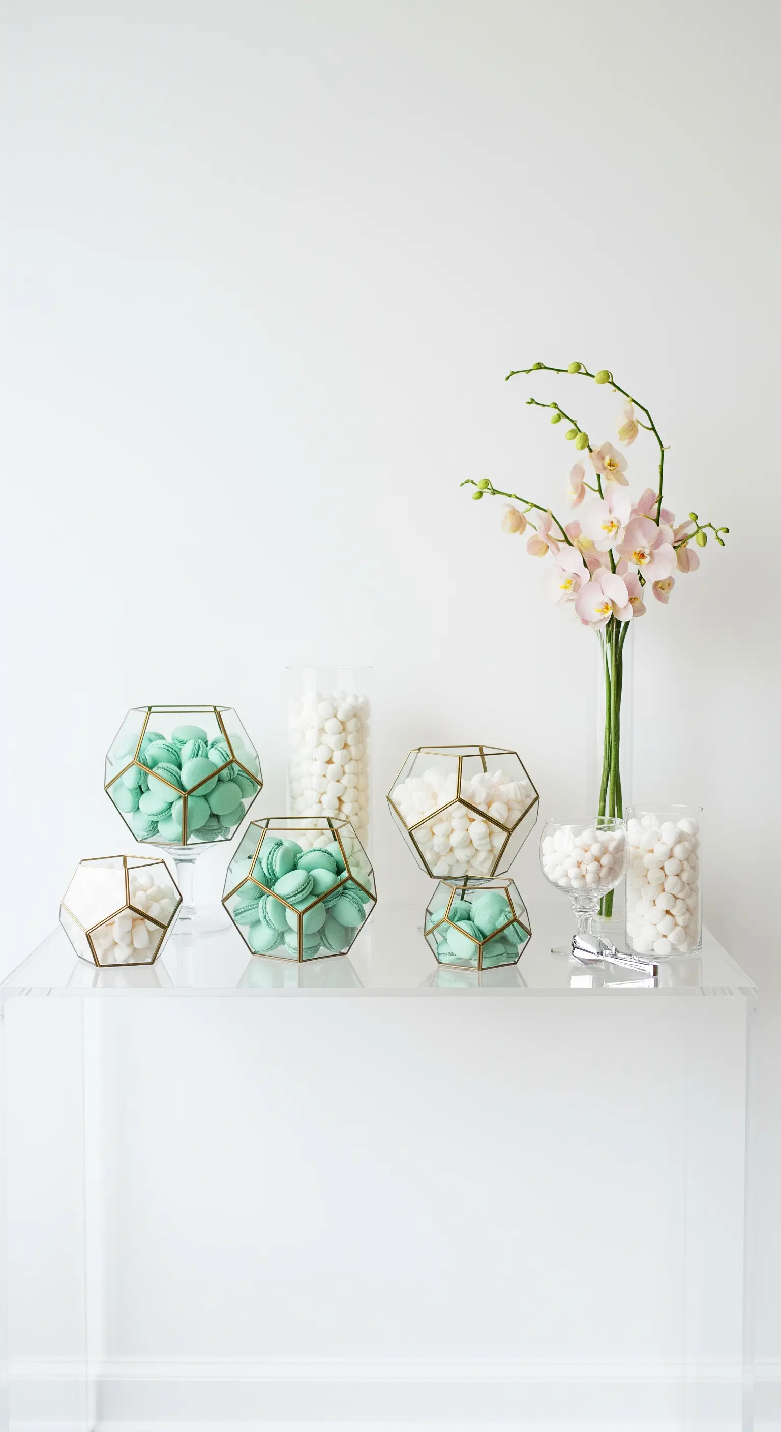 Macarons e marshmallow verde menta in contenitori geometrici dorati su un tavolo trasparente.