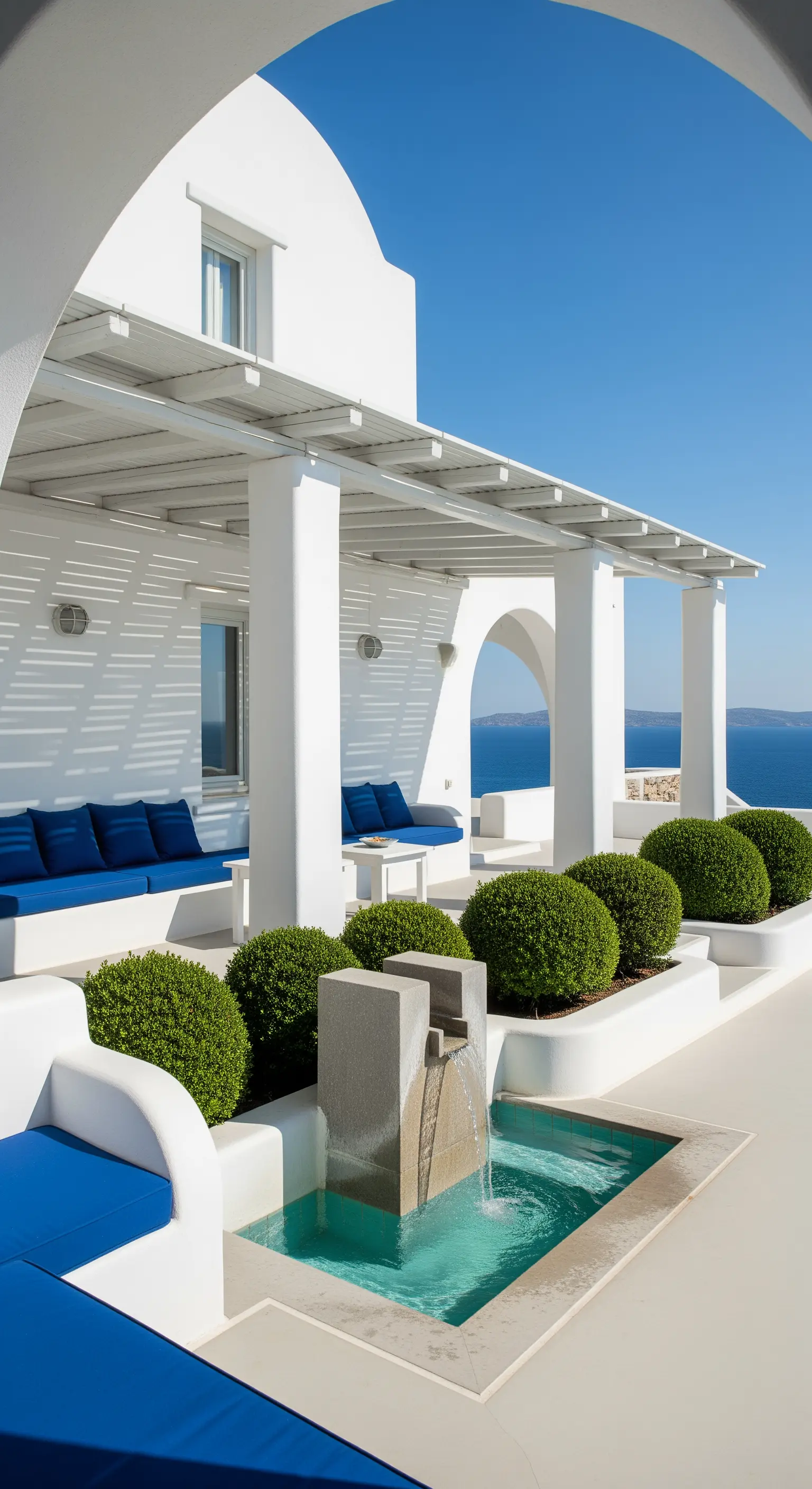 Terrazza panoramica sul mare in stile greco, con arredi bianchi, cuscini blu e siepi sferiche.