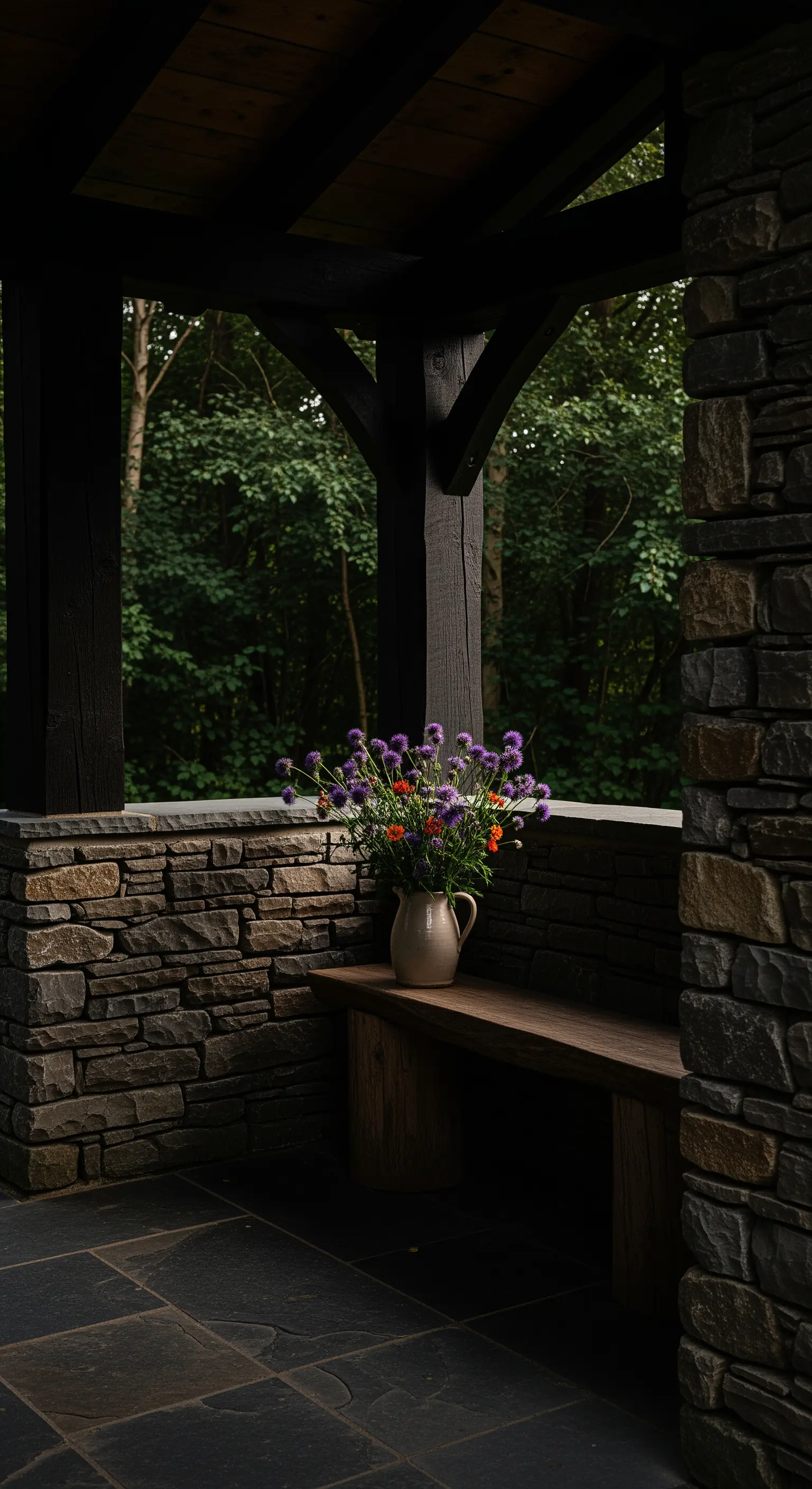 Angolo di un portico scuro con pareti in pietra, panca e un vaso di fiori di campo viola.
