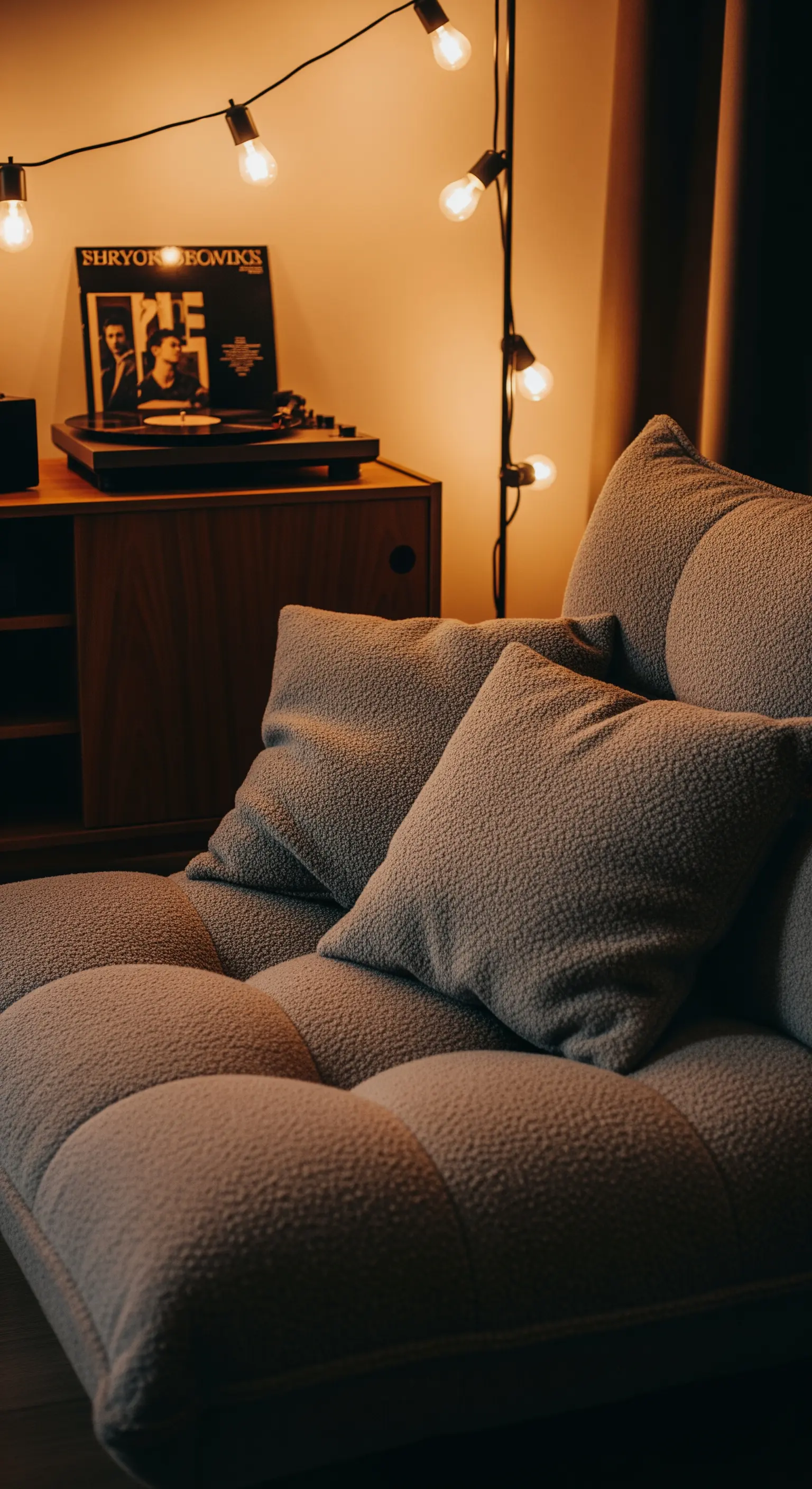Divano basso in tessuto bouclé accanto a un giradischi, con una stringa di luci calde sulla parete.