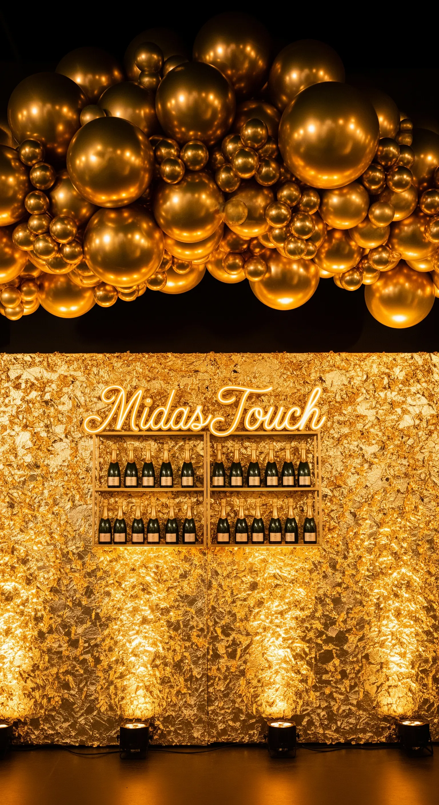Parete rivestita in foglia d'oro con scritta al neon e una nuvola di palloncini dorati.