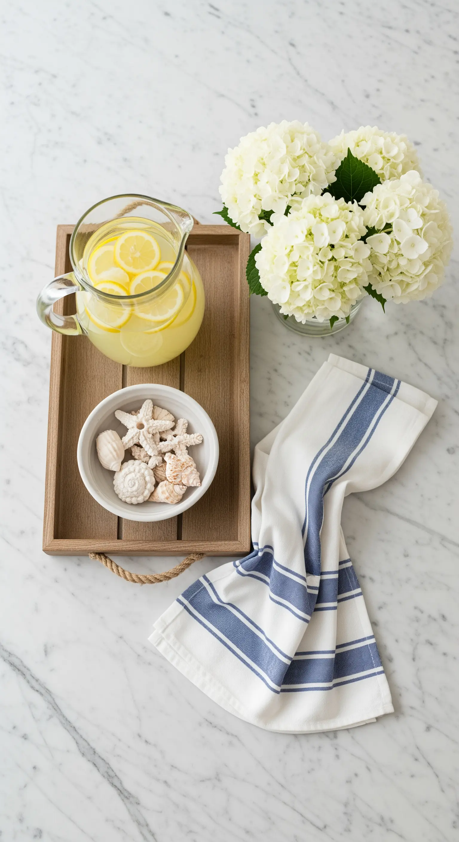 Vassoio in legno con limonata, fiori bianchi e una ciotola di conchiglie su un piano di marmo.