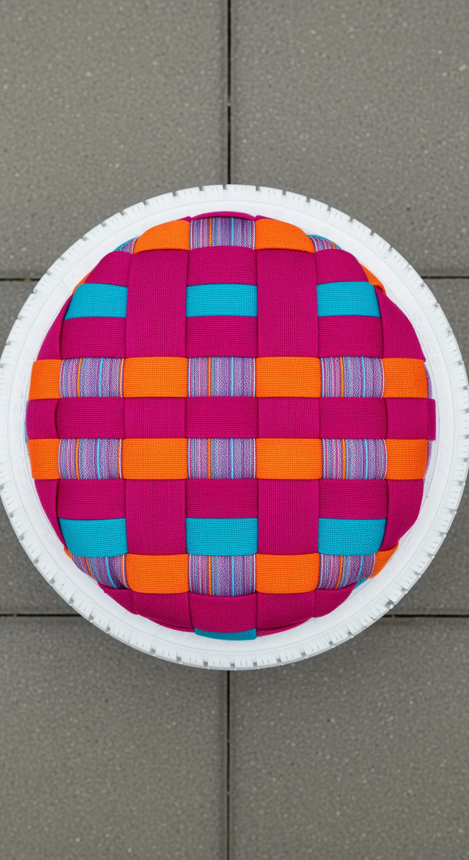 Vista dall'alto di un pouf con seduta realizzata intrecciando cinghie tessili fucsia, arancioni e blu.