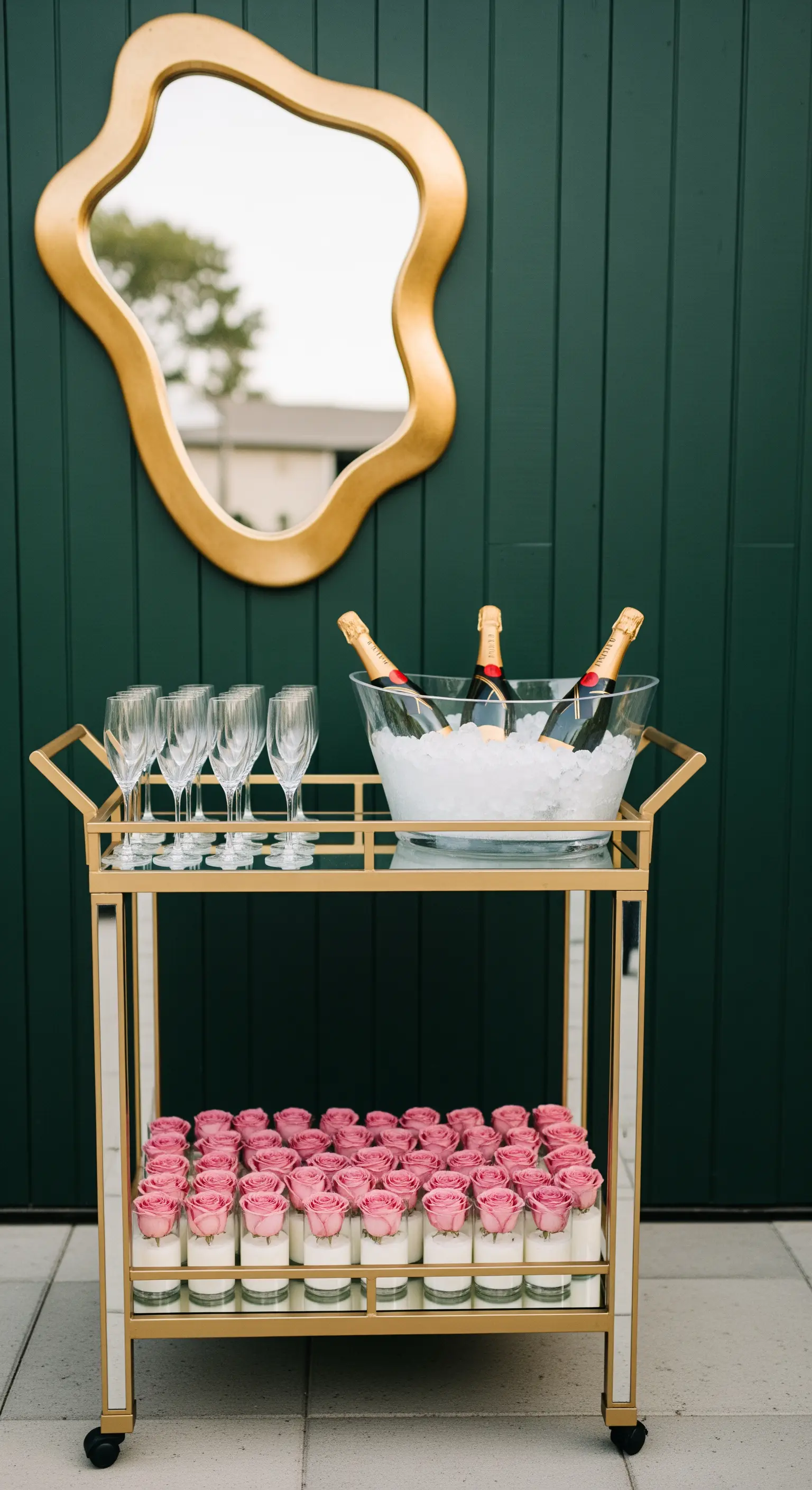 Carrello bar dorato con champagne e candele, davanti a una parete verde con uno specchio organico.