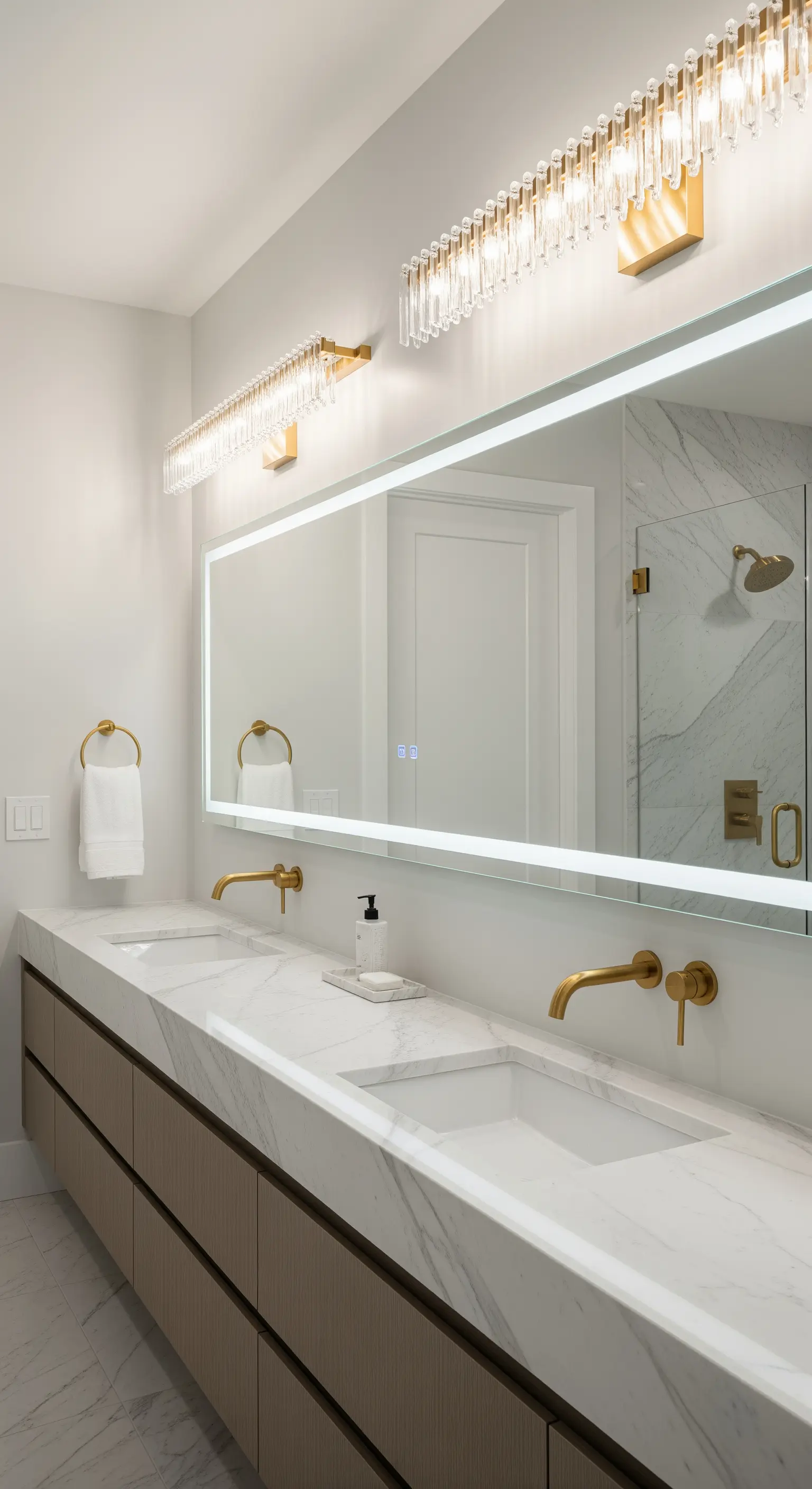 Lungo specchio da bagno con retroilluminazione a LED sopra un doppio lavabo in marmo con rubinetti dorati.