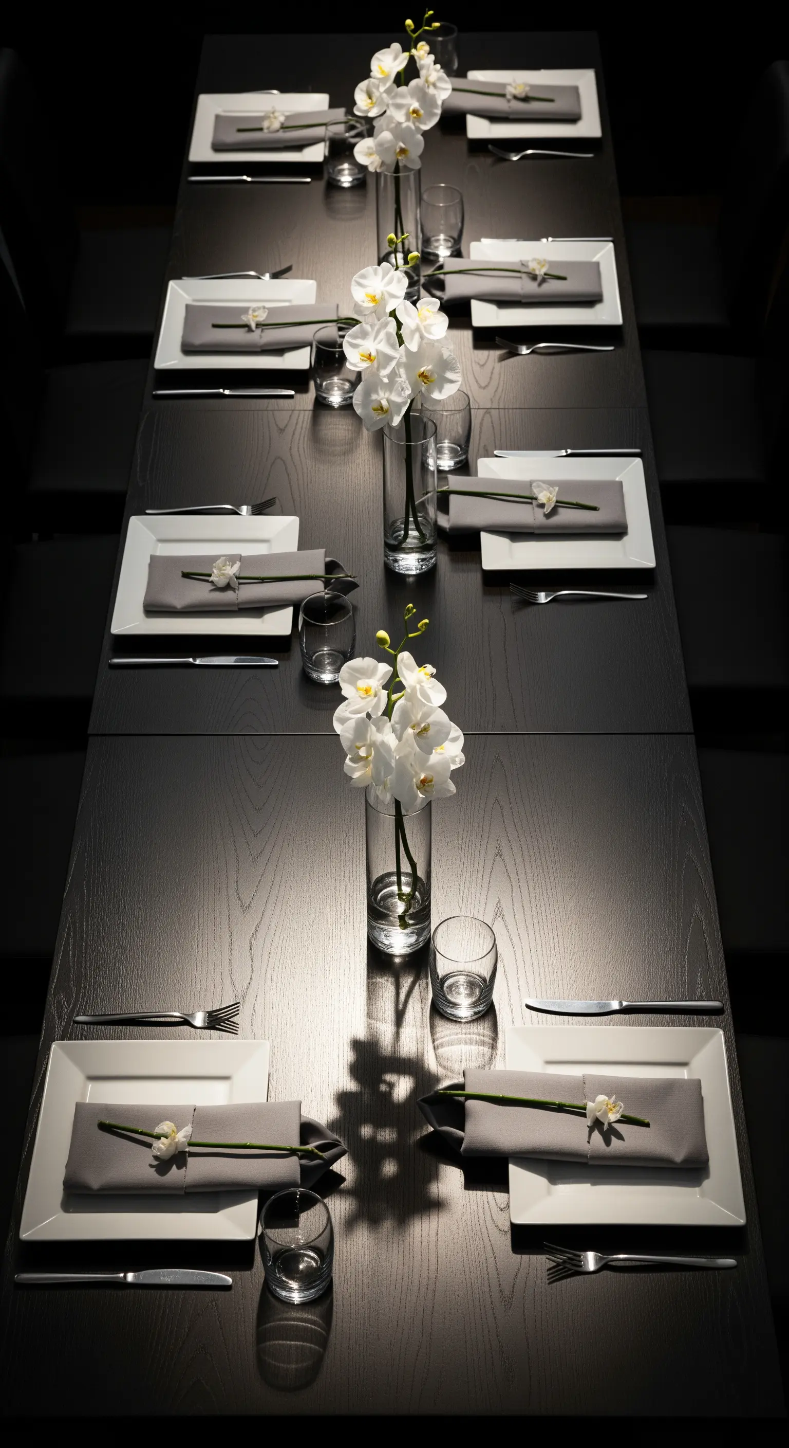 Lunga tavola scura illuminata da faretti, con vasi di orchidee bianche.