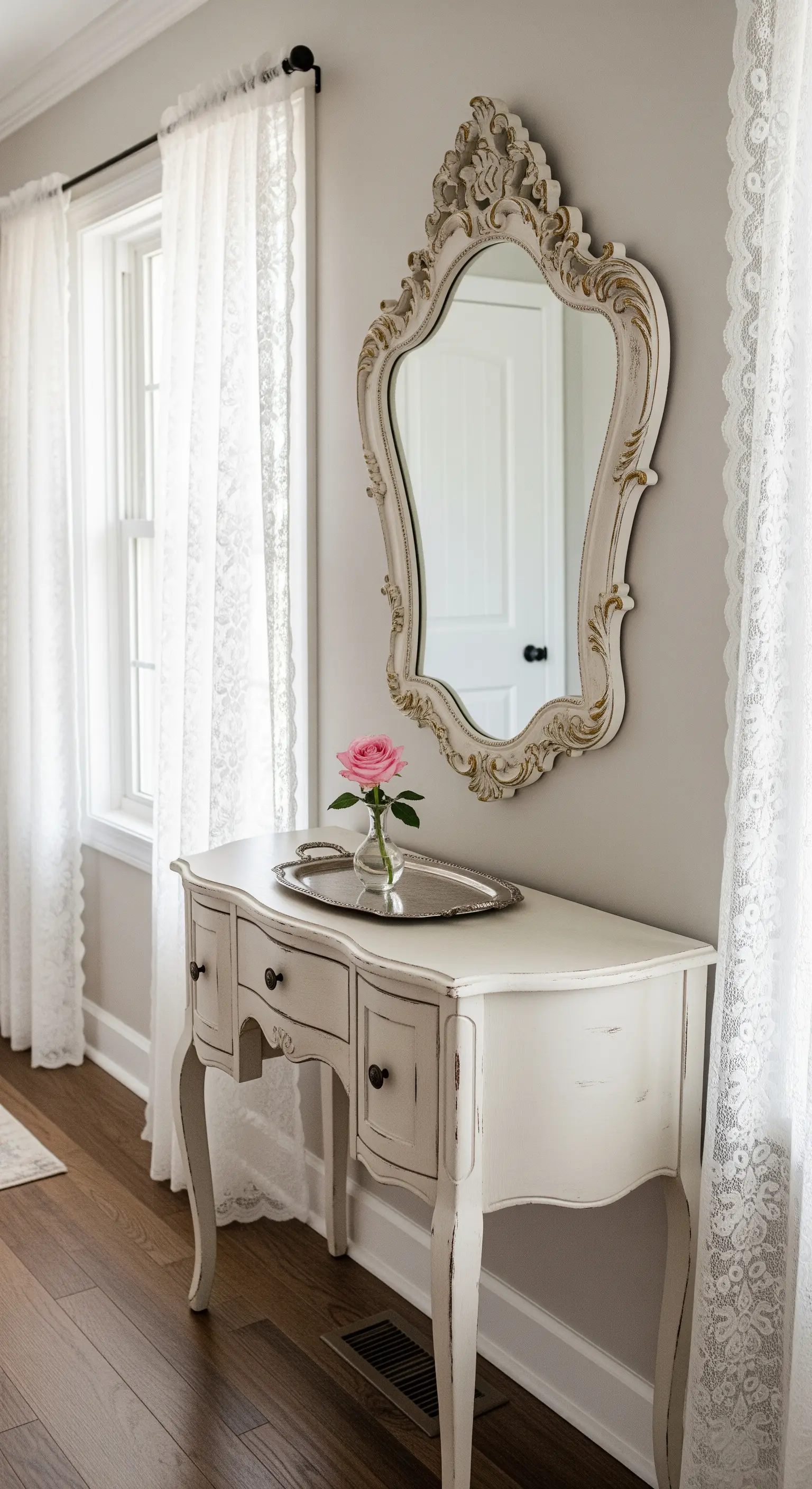 Consolle bianca con specchio barocco, incorniciata da tende di pizzo bianco che filtrano la luce.