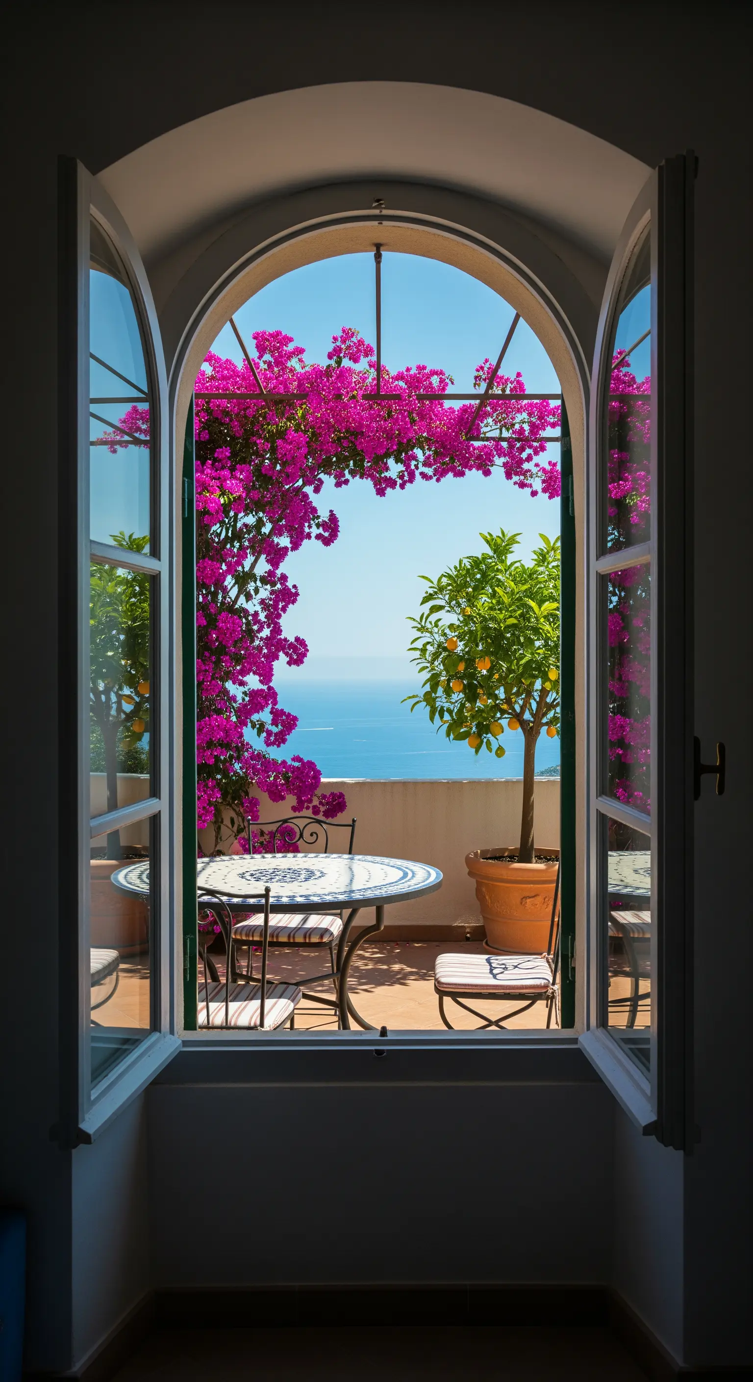 Vista da una finestra su un balcone con bouganville, tavolo in mosaico e mare.