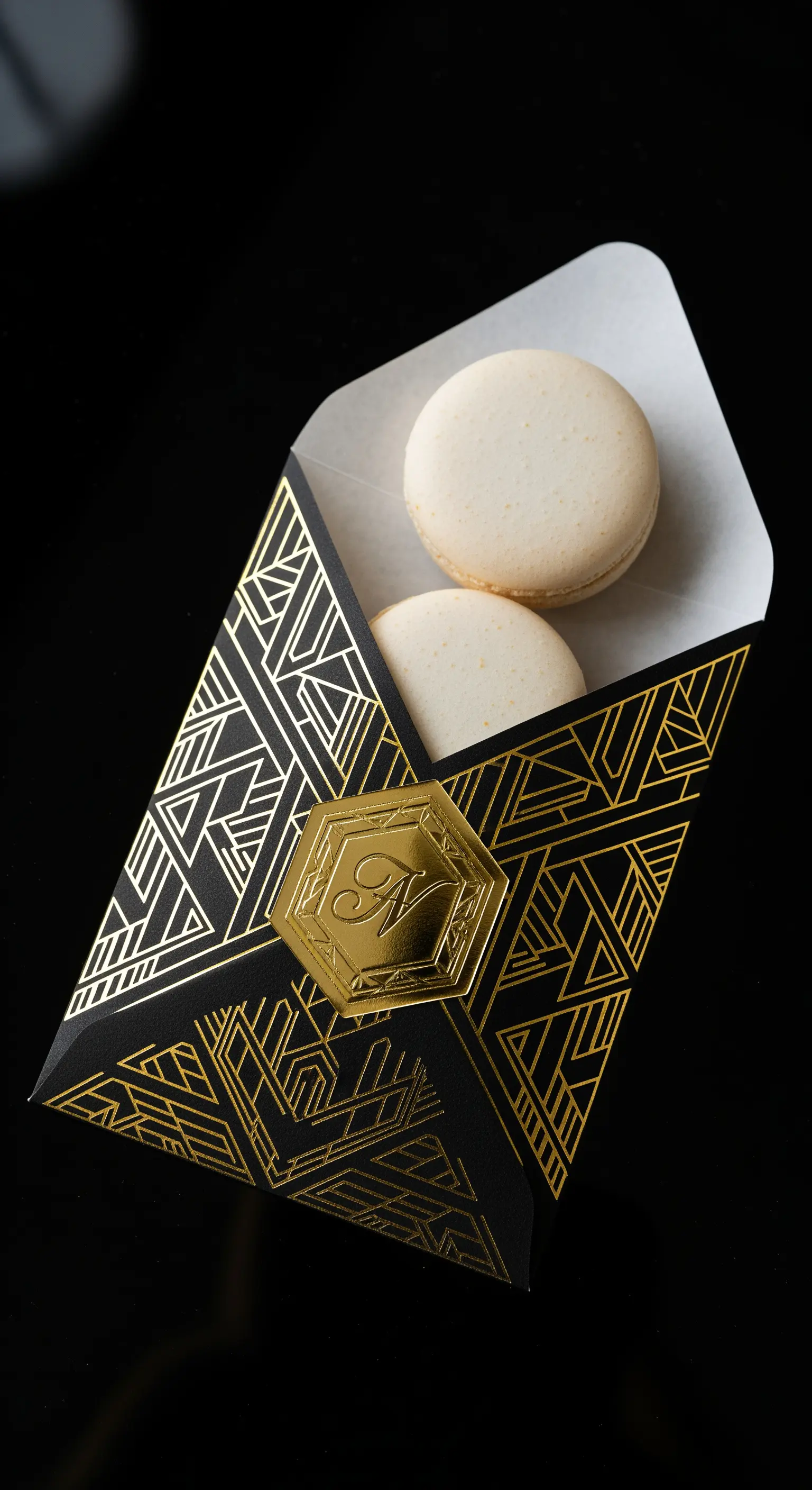 Busta nera con motivi geometrici dorati in stile Art Déco, contenente due macarons bianchi.
