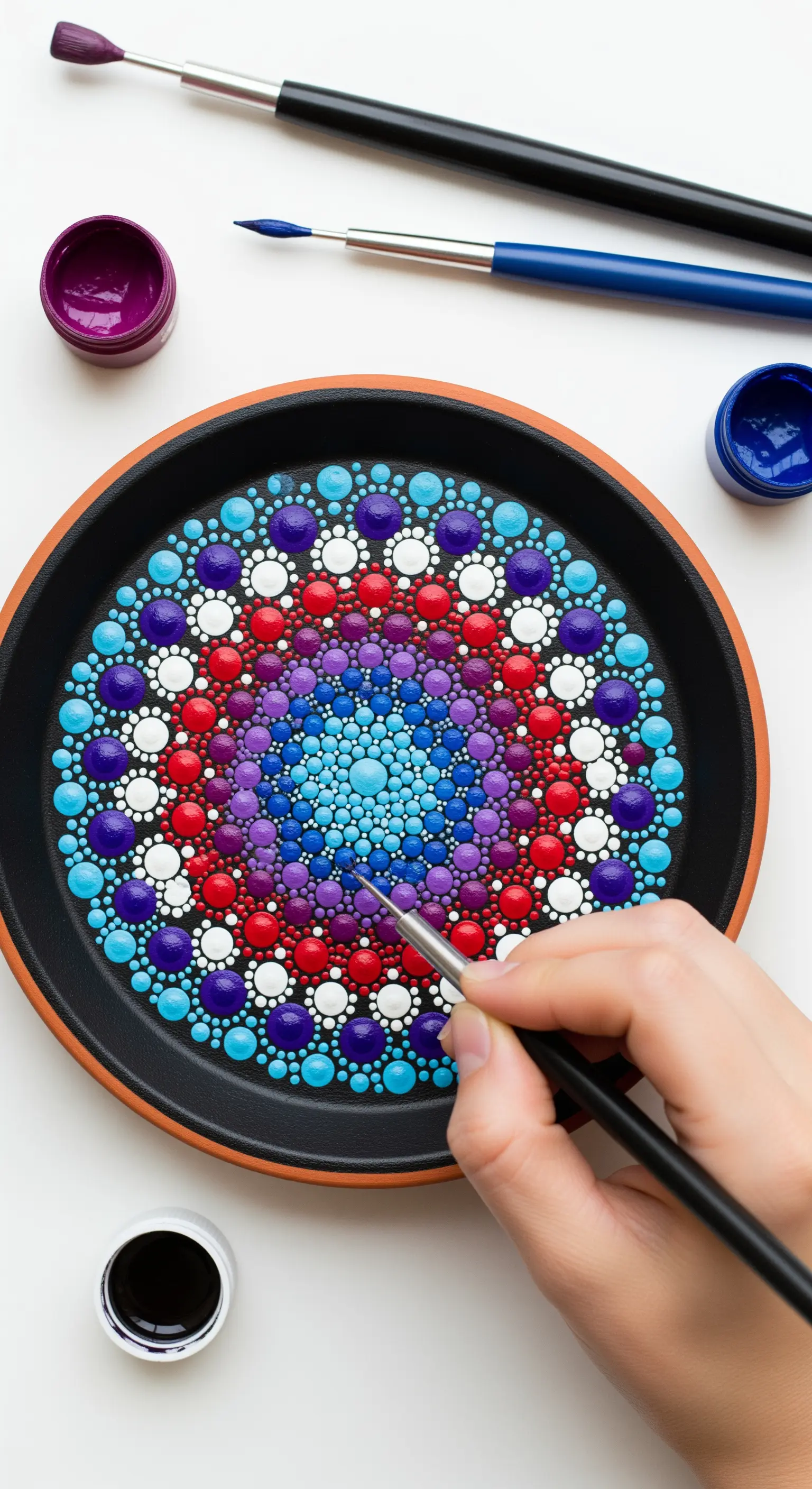 Mano che dipinge un mandala di punti colorati su un sottovaso nero usando un pennello sottile.