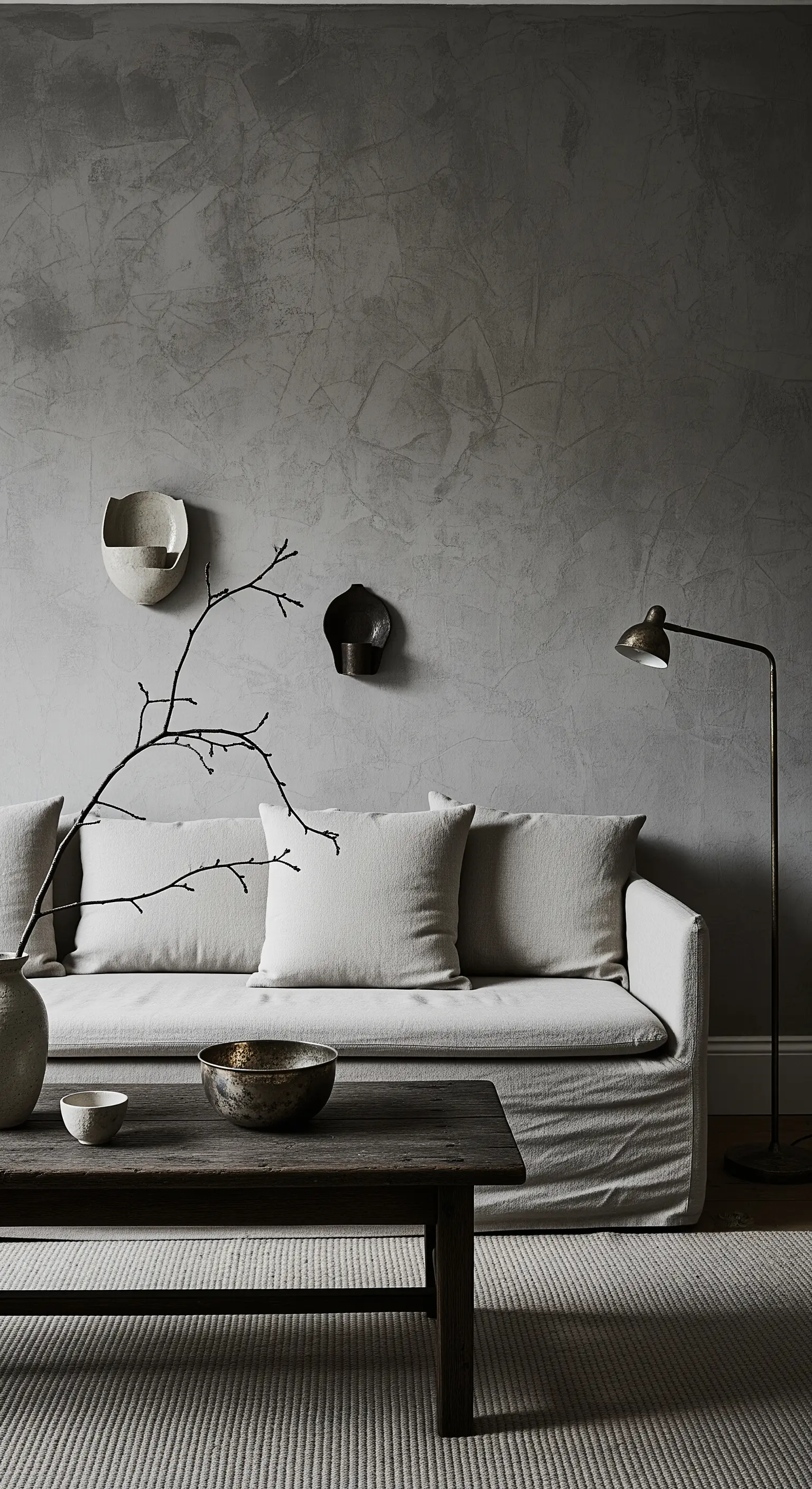 Salotto minimalista con parete grigia materica, divano in lino chiaro e dettagli scuri.