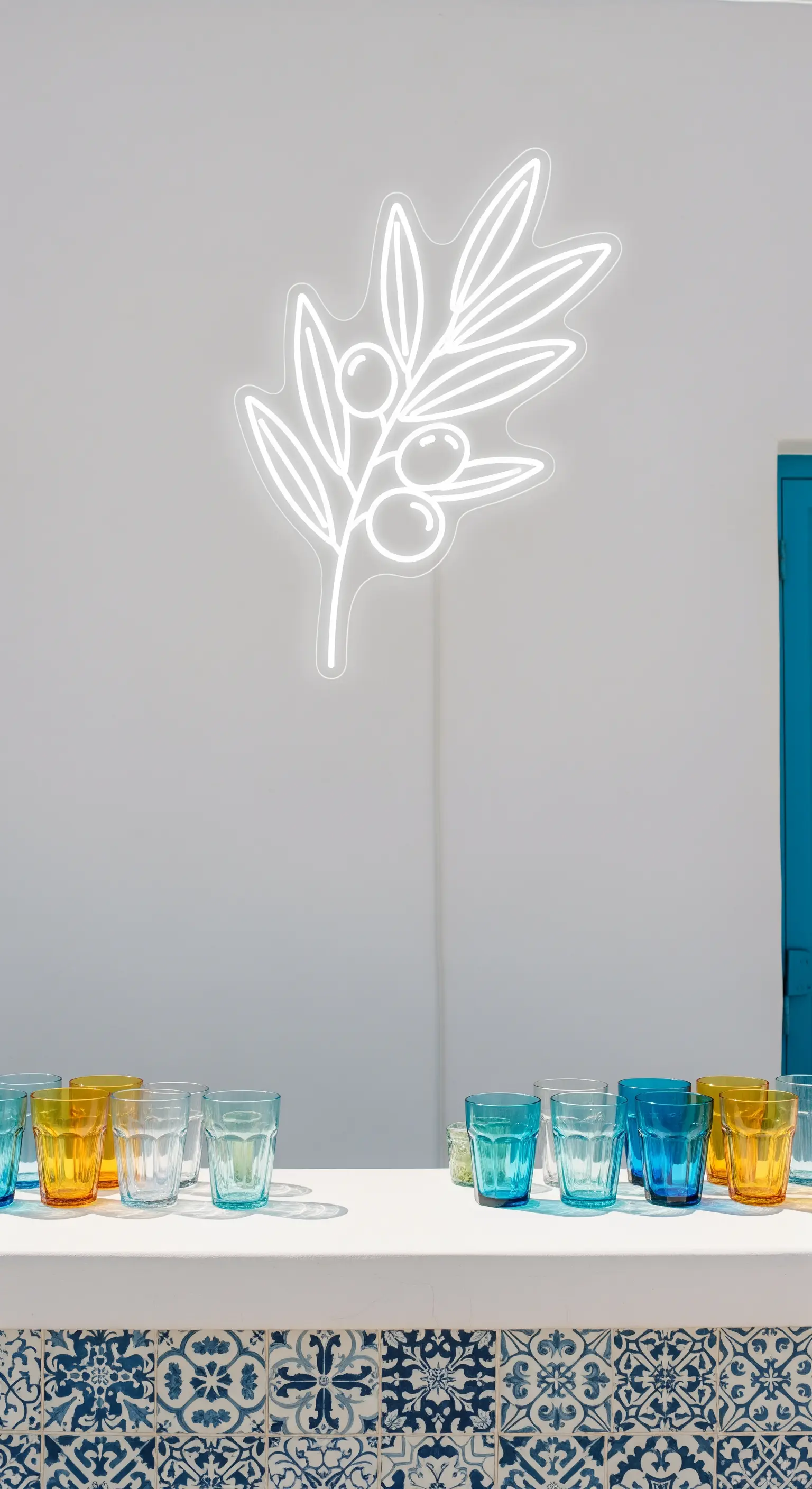 Neon bianco a forma di ramo d'ulivo su una parete bianca, con bar rivestito di maioliche blu.