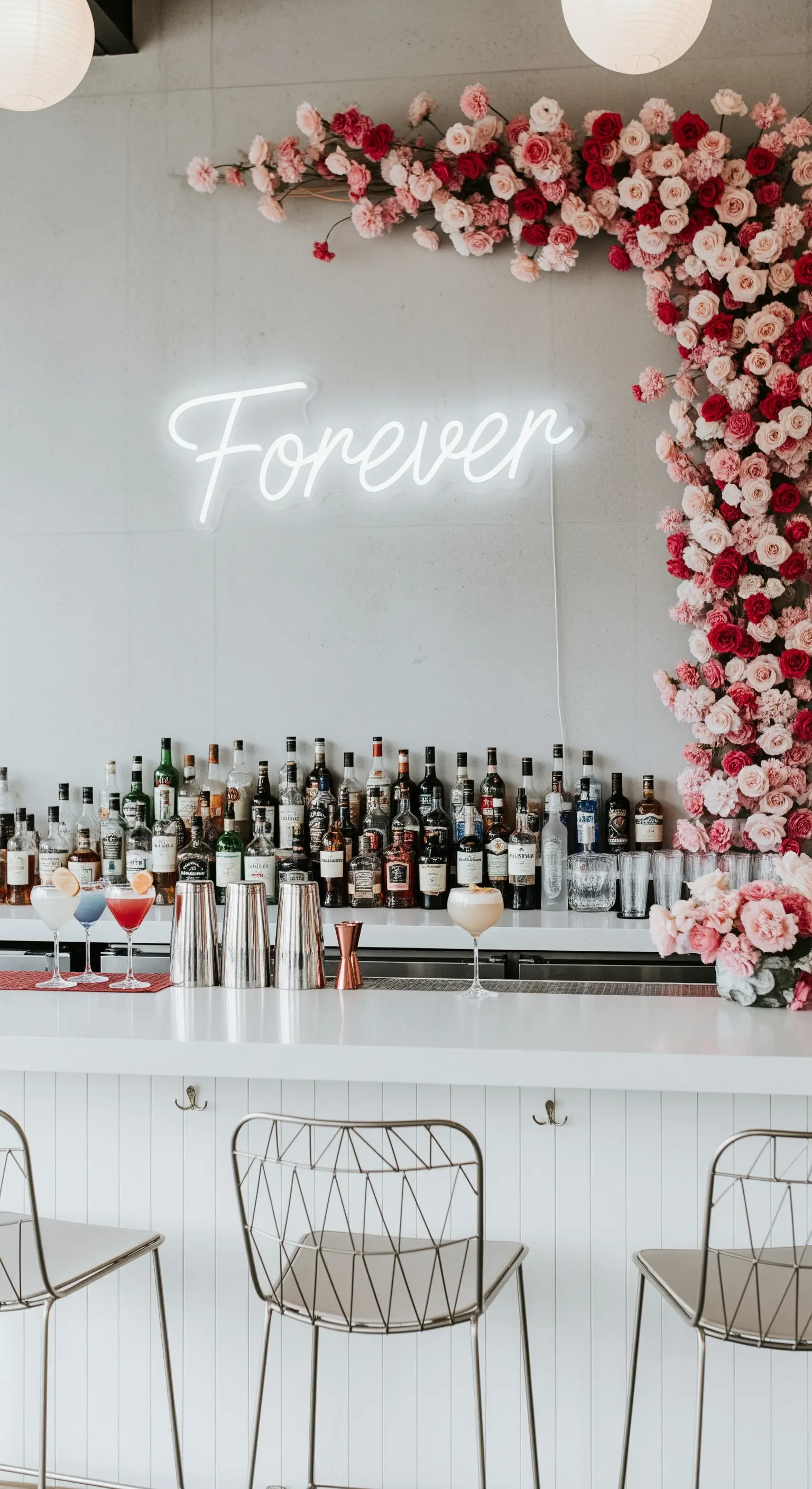 Scritta al neon bianca “Forever” su una parete bianca, con un angolo decorato da rose rosa.