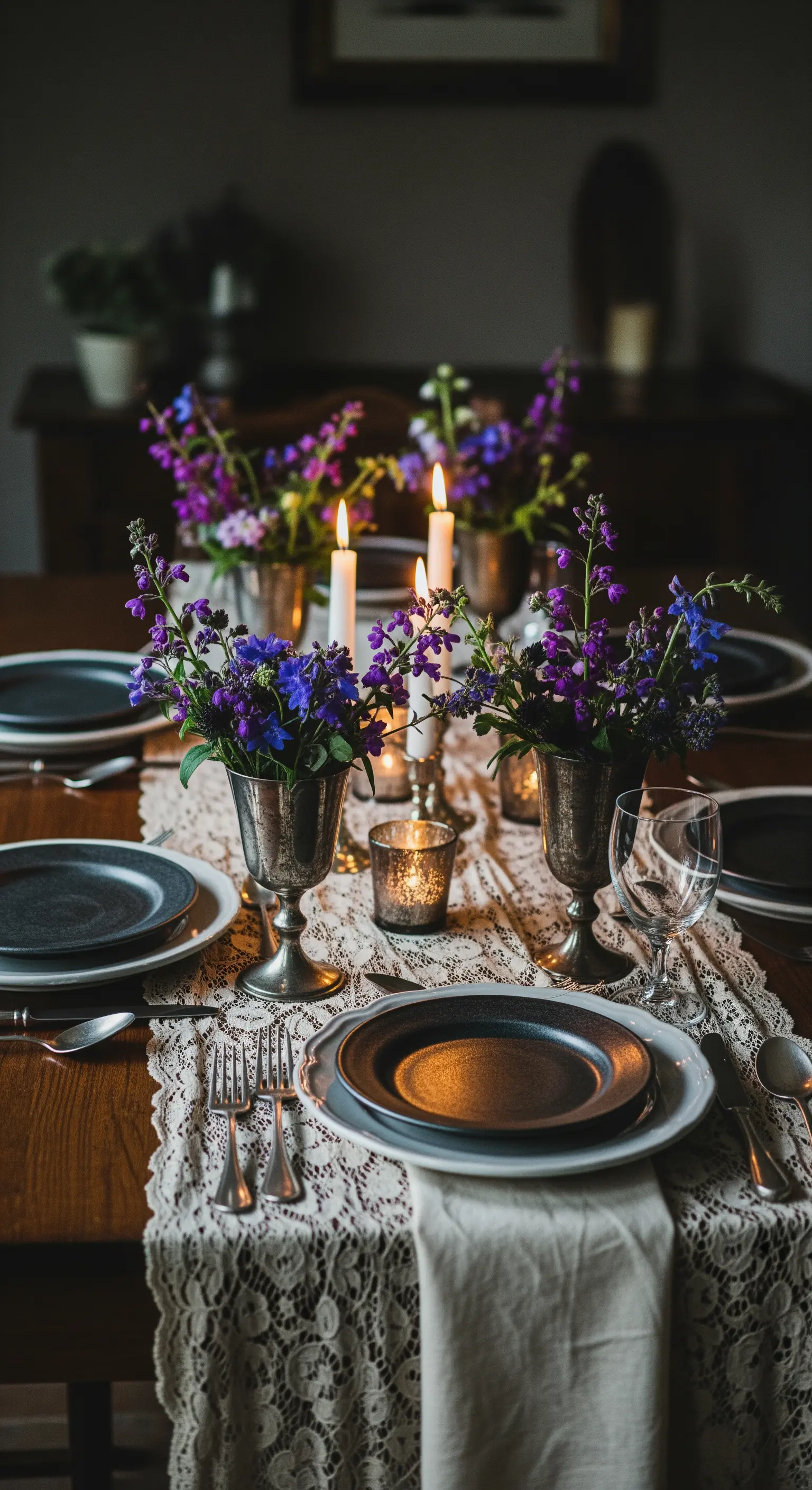 Tavola scura illuminata da candele, con runner in pizzo, piatti neri e coppe d'argento