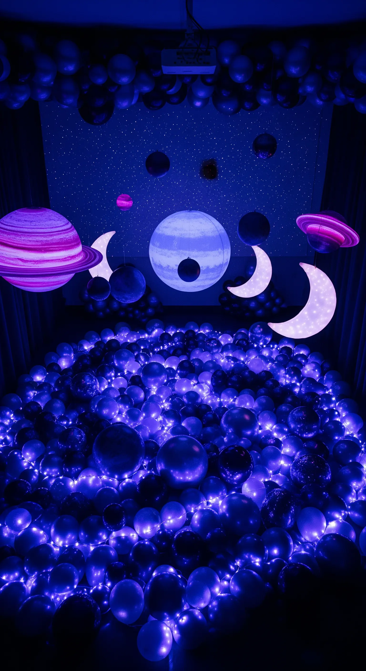 Stanza buia piena di palloncini blu e viola illuminati da lucine, con pianeti sospesi.