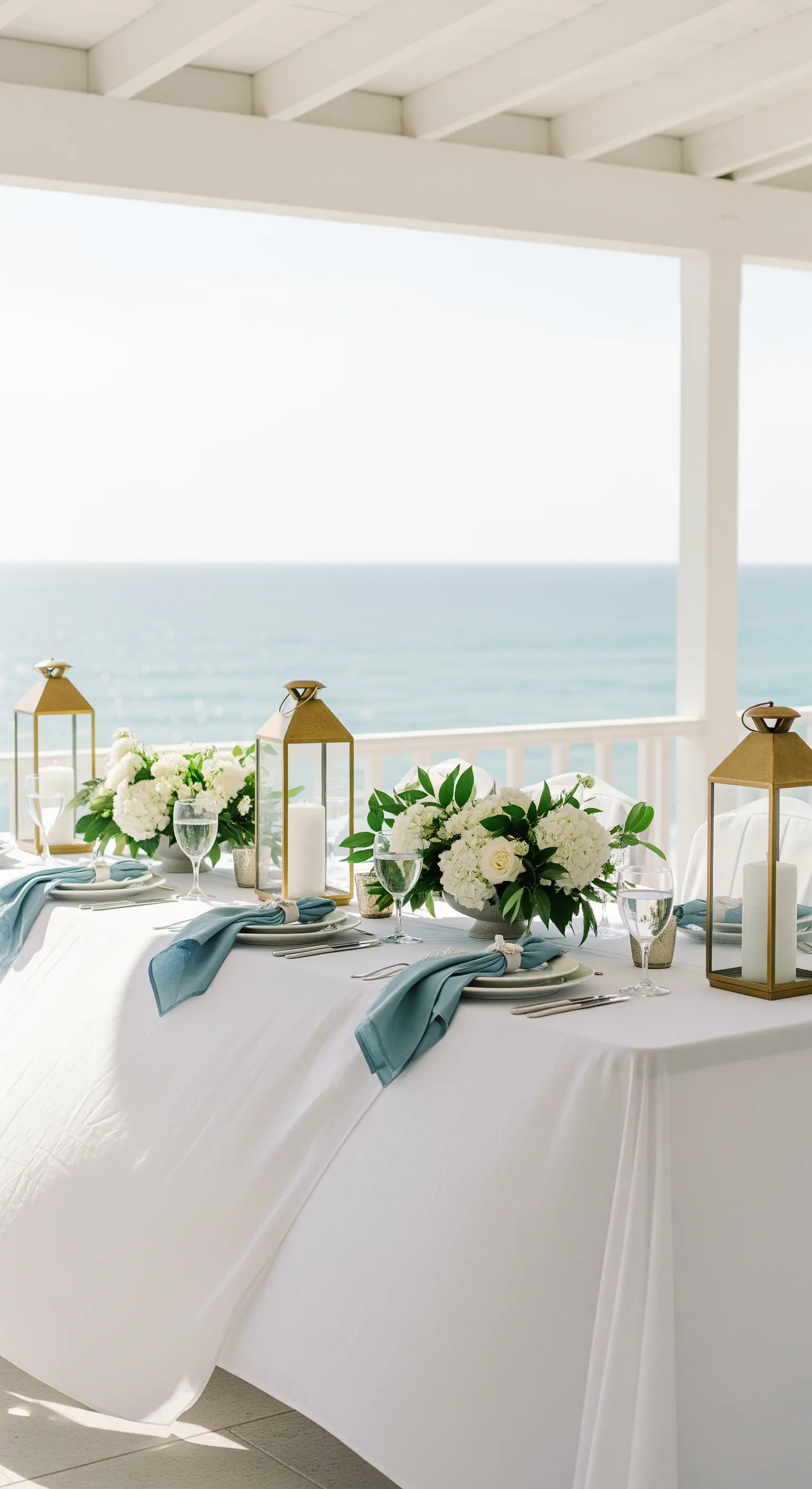Tavola vista mare con lanterne dorate, tovaglioli azzurri e ortensie bianche.