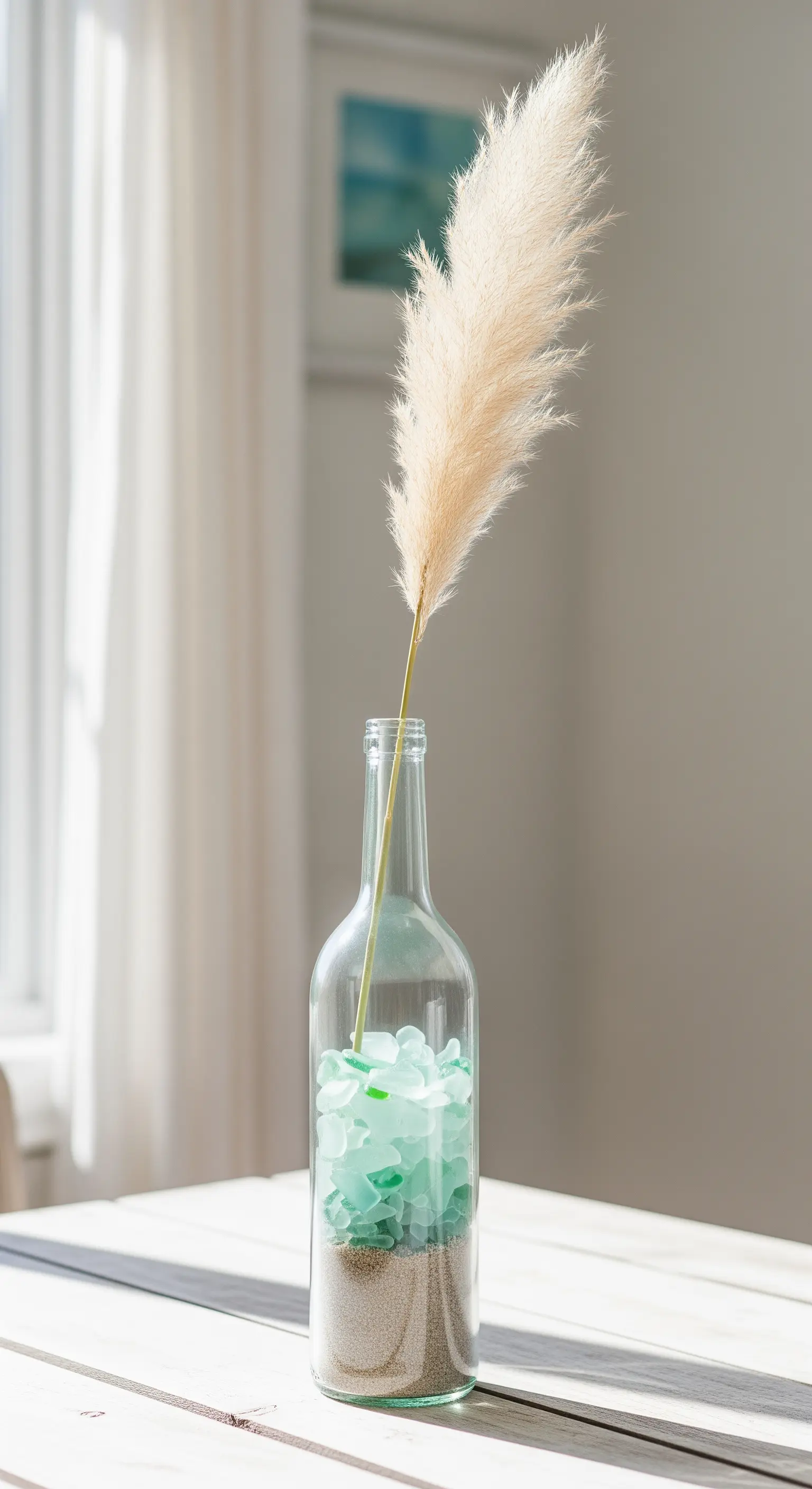Bottiglia di vino trasparente riempita con sabbia, vetri marini e un ramo di pampa.