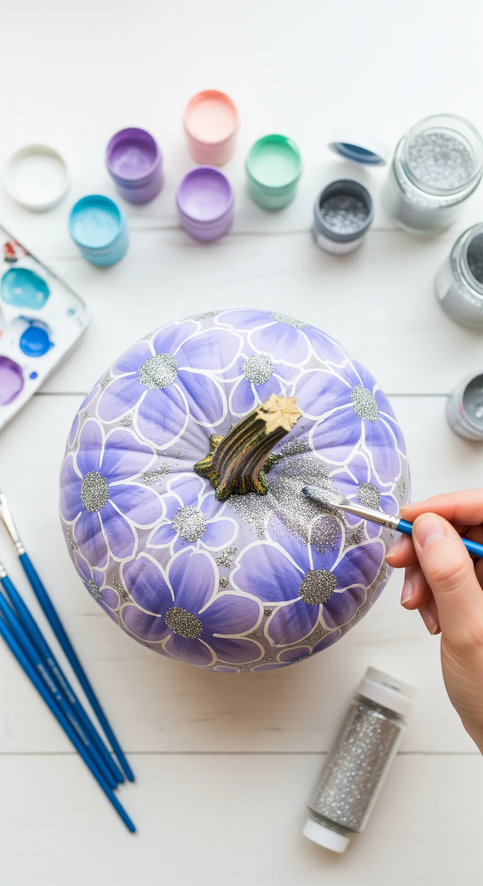 Mano che dipinge fiori viola su una zucca, aggiungendo glitter argento al centro.
