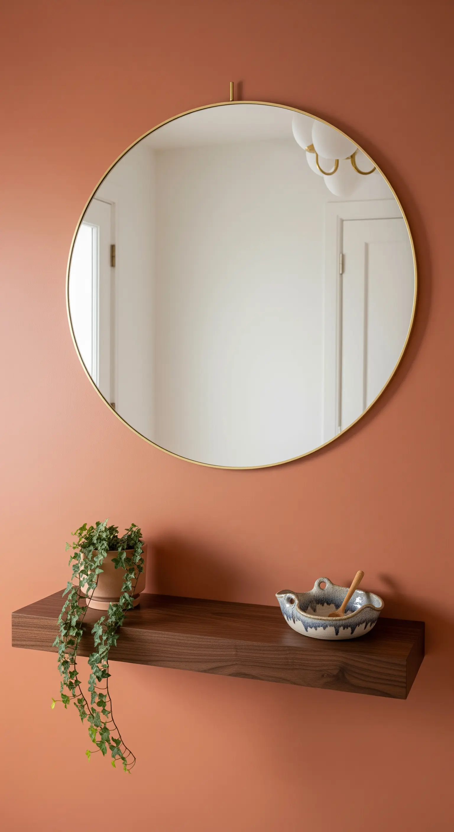 Mensola sospesa in legno con specchio rotondo e una piccola pianta, su una parete terracotta.