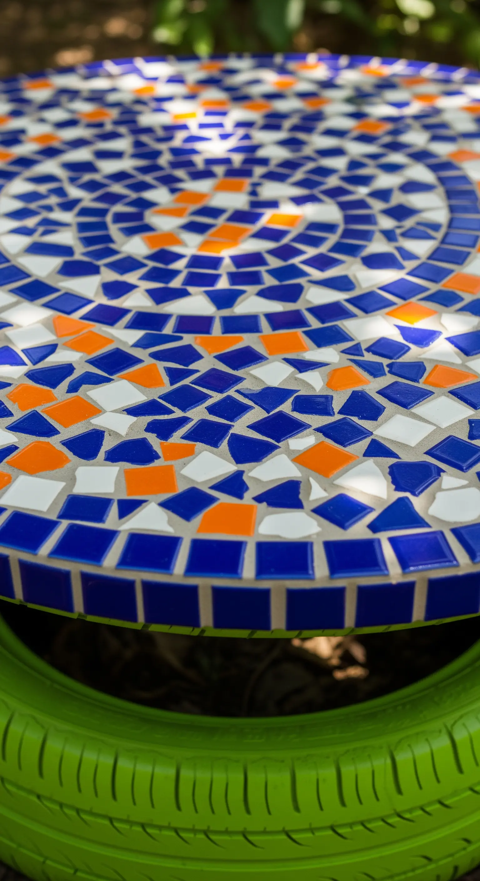 Tavolino da giardino con base in pneumatico verde e piano decorato con mosaico blu, bianco e arancio.