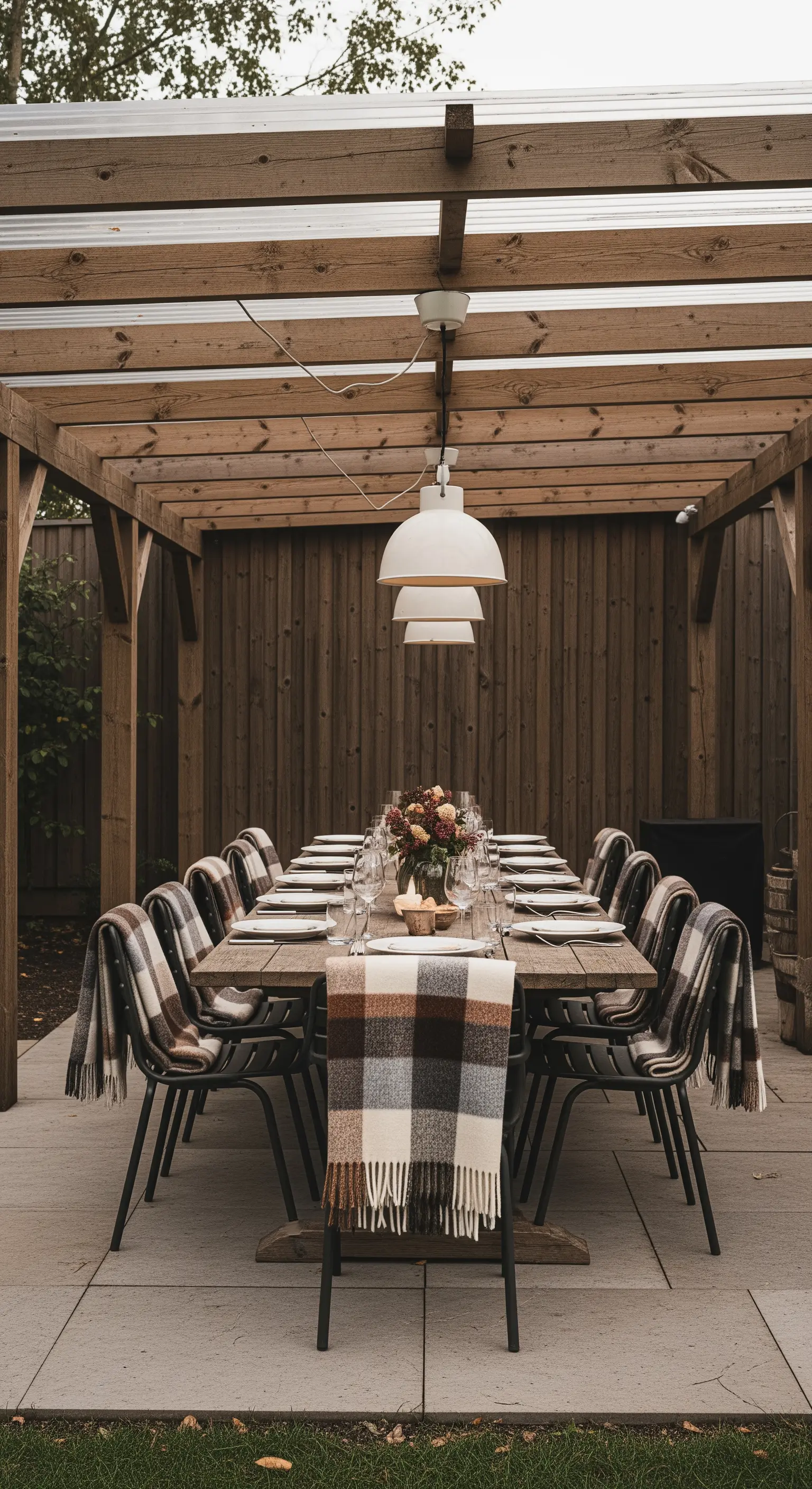 Lungo tavolo da pranzo in legno sotto una pergola, con plaid a quadri su ogni sedia.