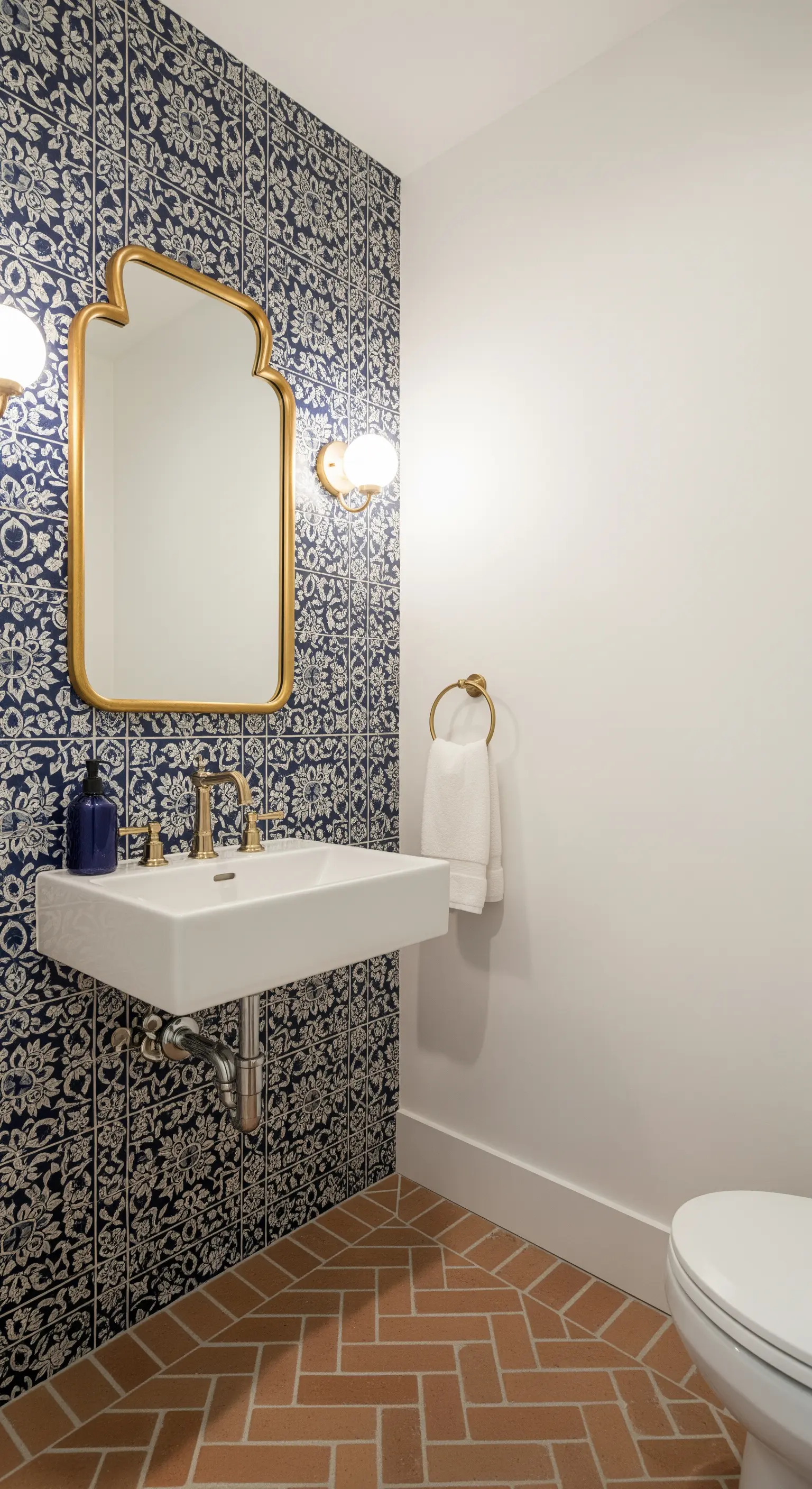 Bagno di servizio con parete a motivi blu, specchio dorato e pavimento in cotto a spina di pesce.