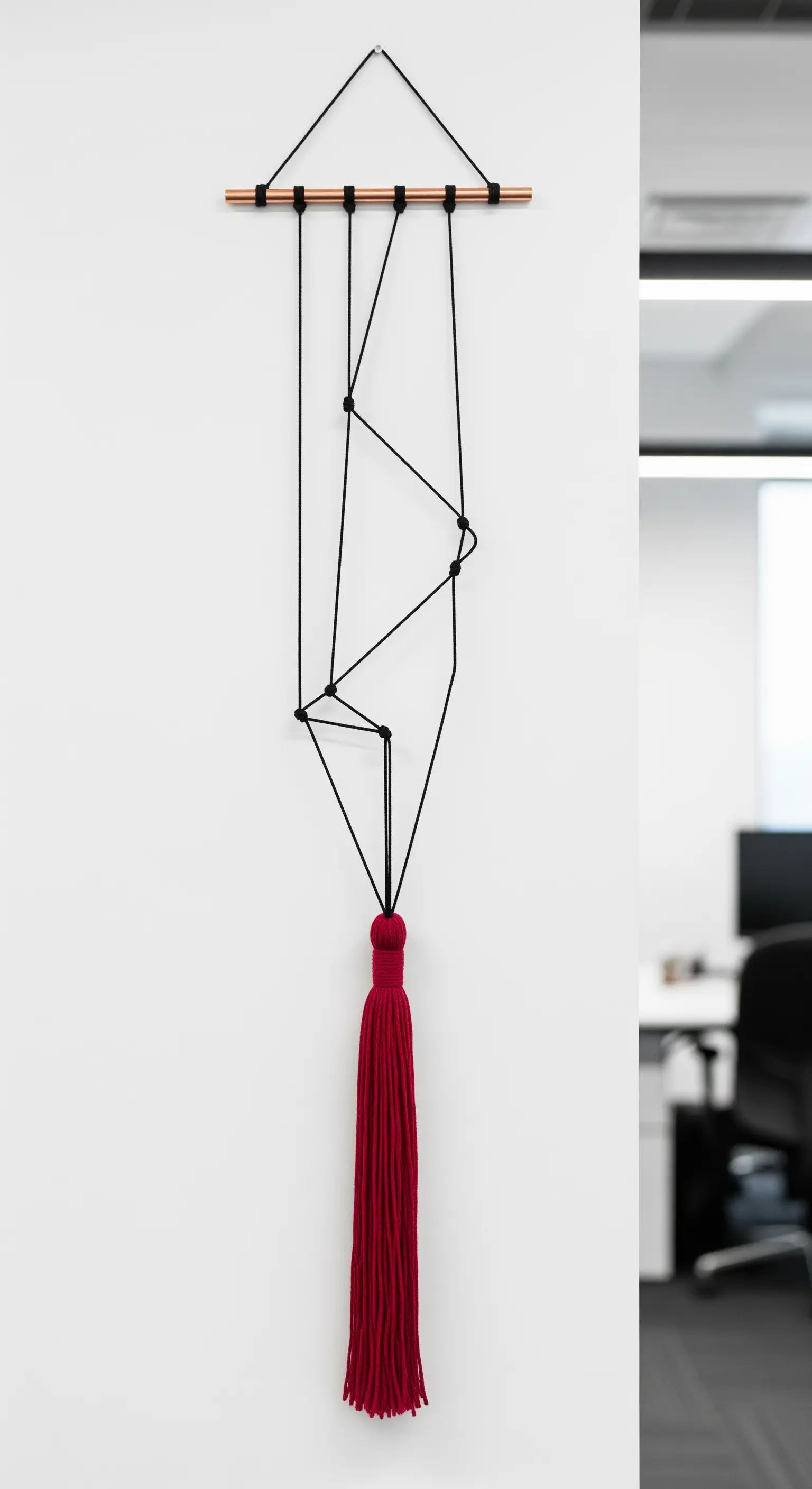 Decorazione murale minimalista con filo nero teso a formare linee astratte e una nappa rossa.