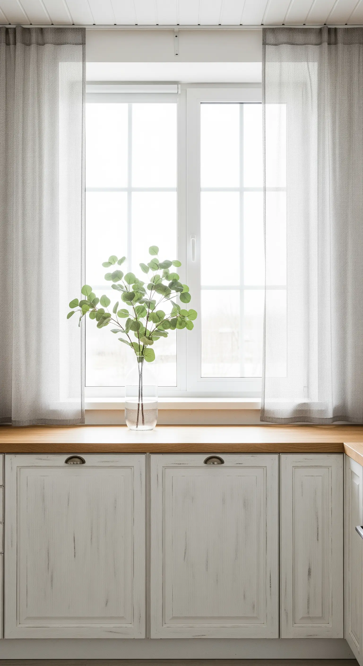 Mobili da cucina bianchi decapati con piano in legno chiaro e tende grigie.