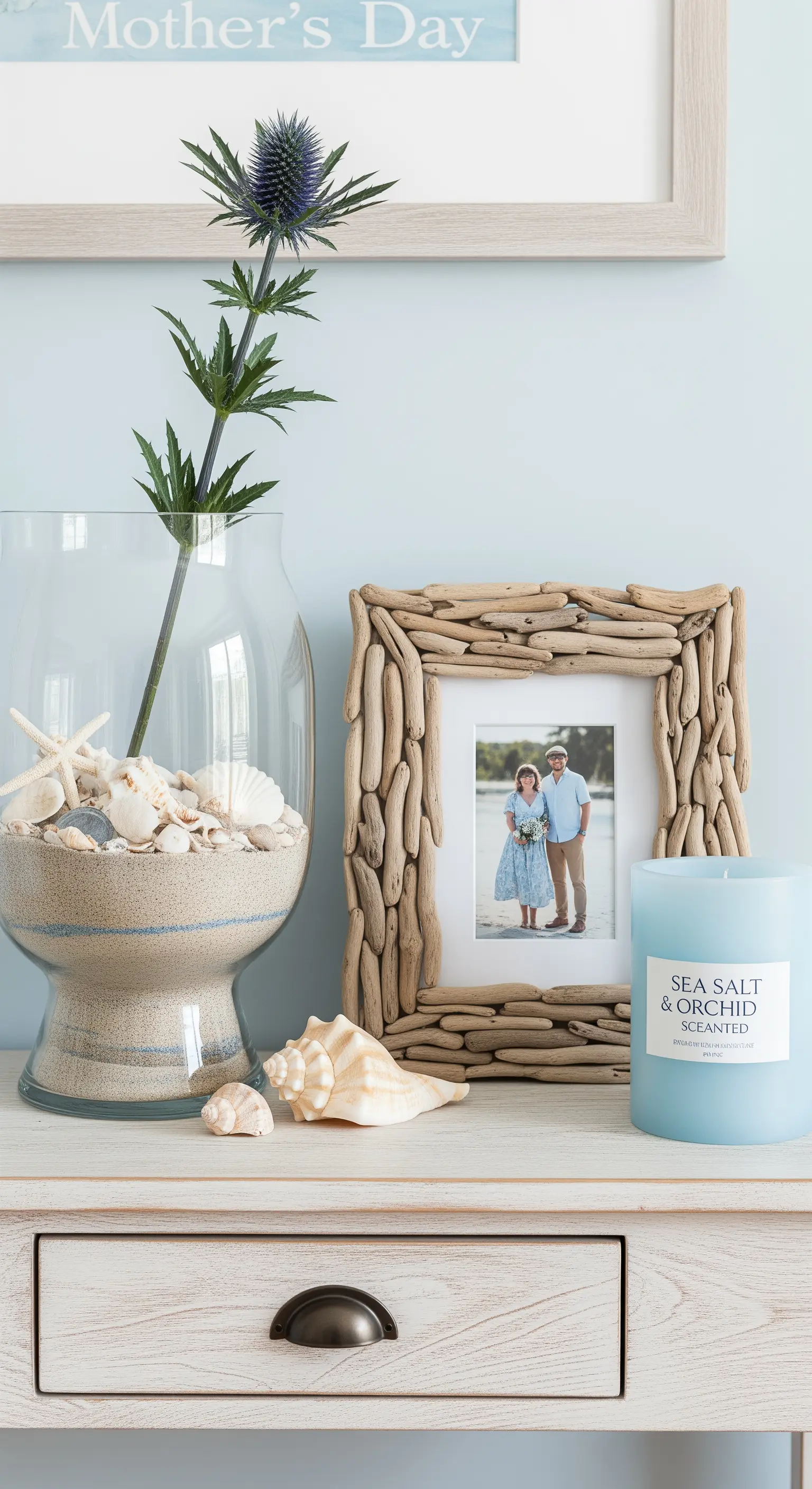 Decorazione a tema marino con cornice di legni, vaso con sabbia e conchiglie, e candela blu.