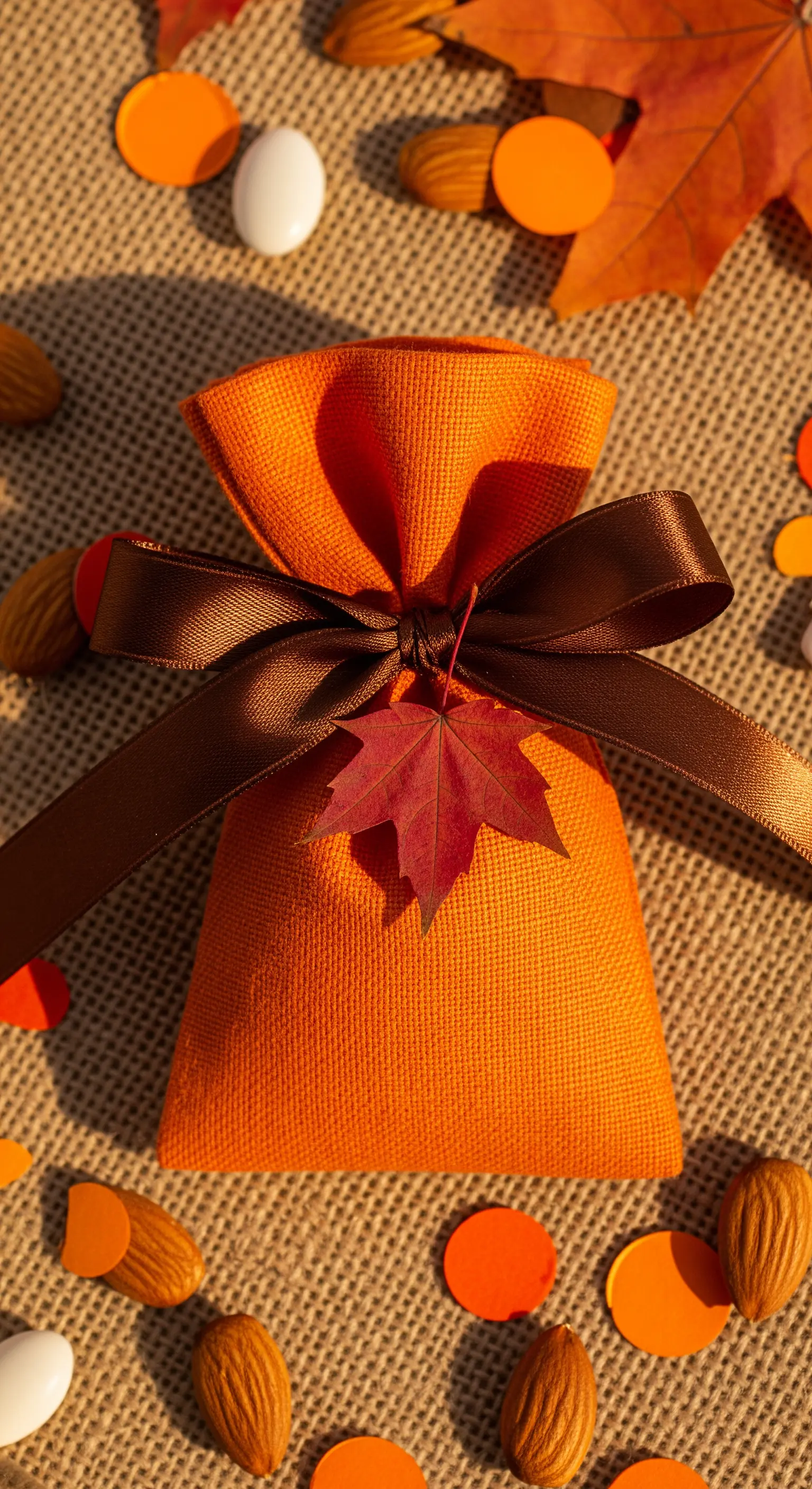 Sacchetto bomboniera arancione con nastro marrone e una foglia d'acero rossa, su un fondo di juta.