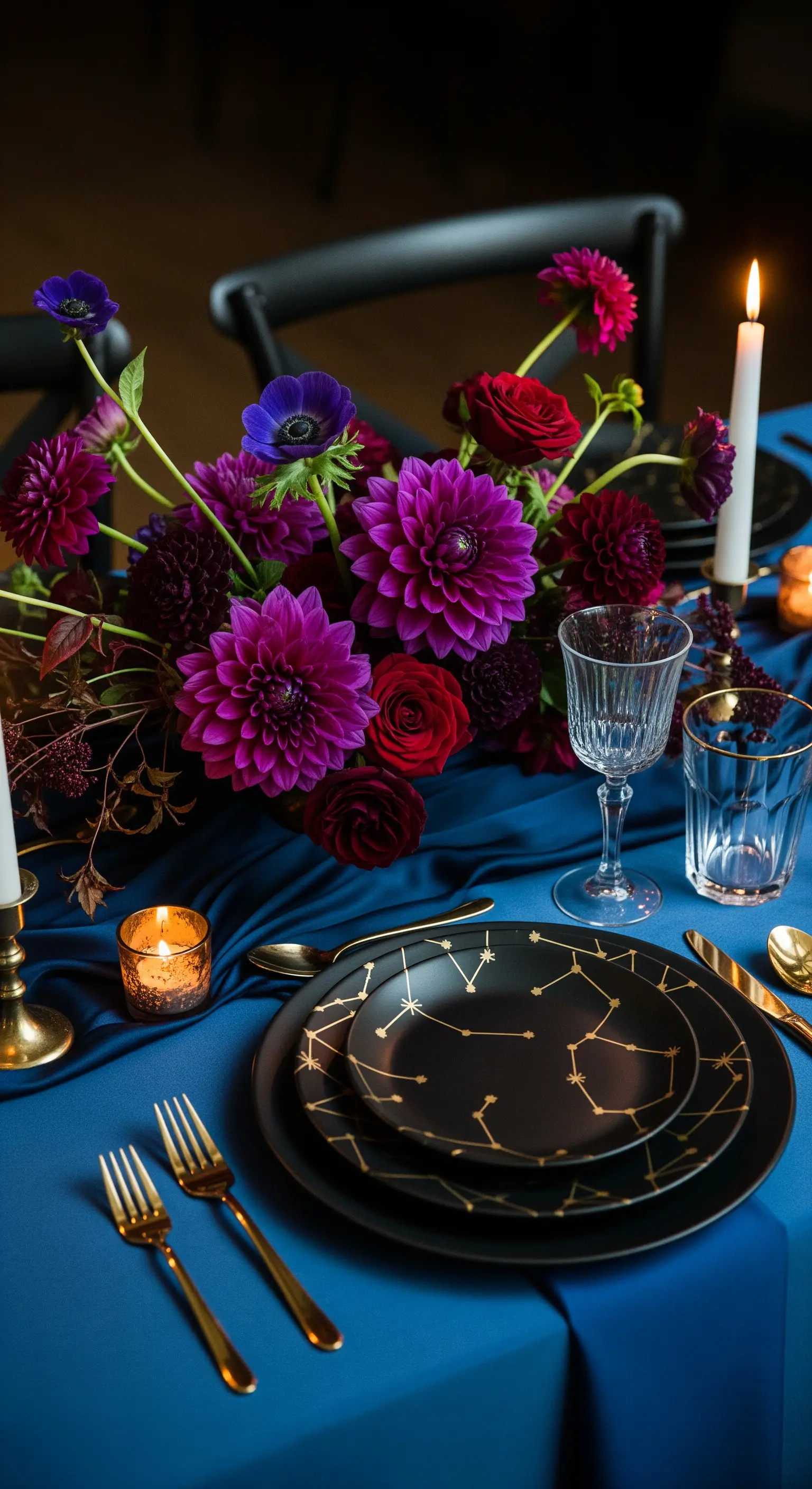 Tavola blu notte con piatti neri a tema costellazioni, posate dorate e un bouquet di fiori scuri.