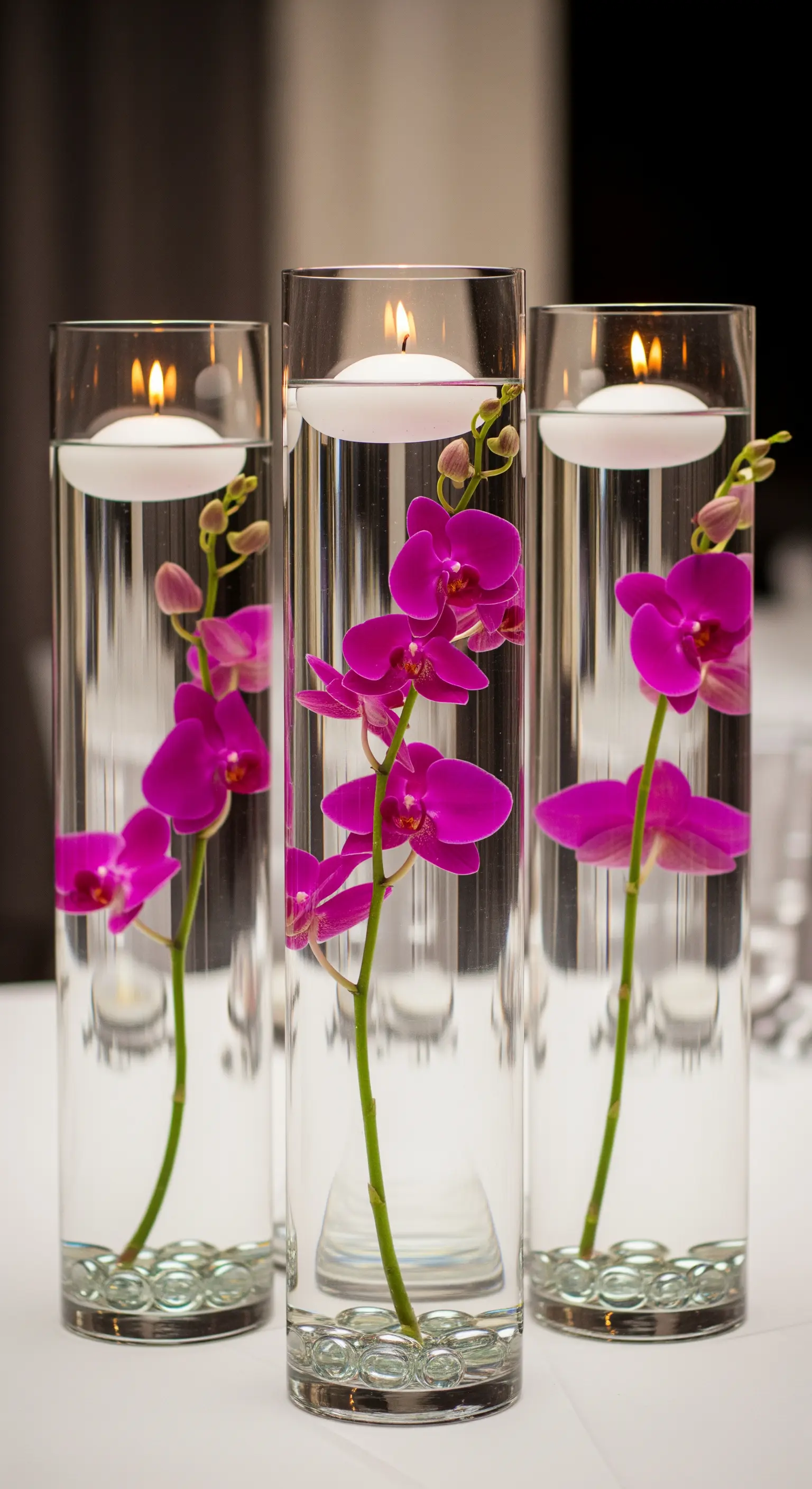 Alti vasi cilindrici con steli di orchidea fucsia immersi nell'acqua e candele galleggianti.