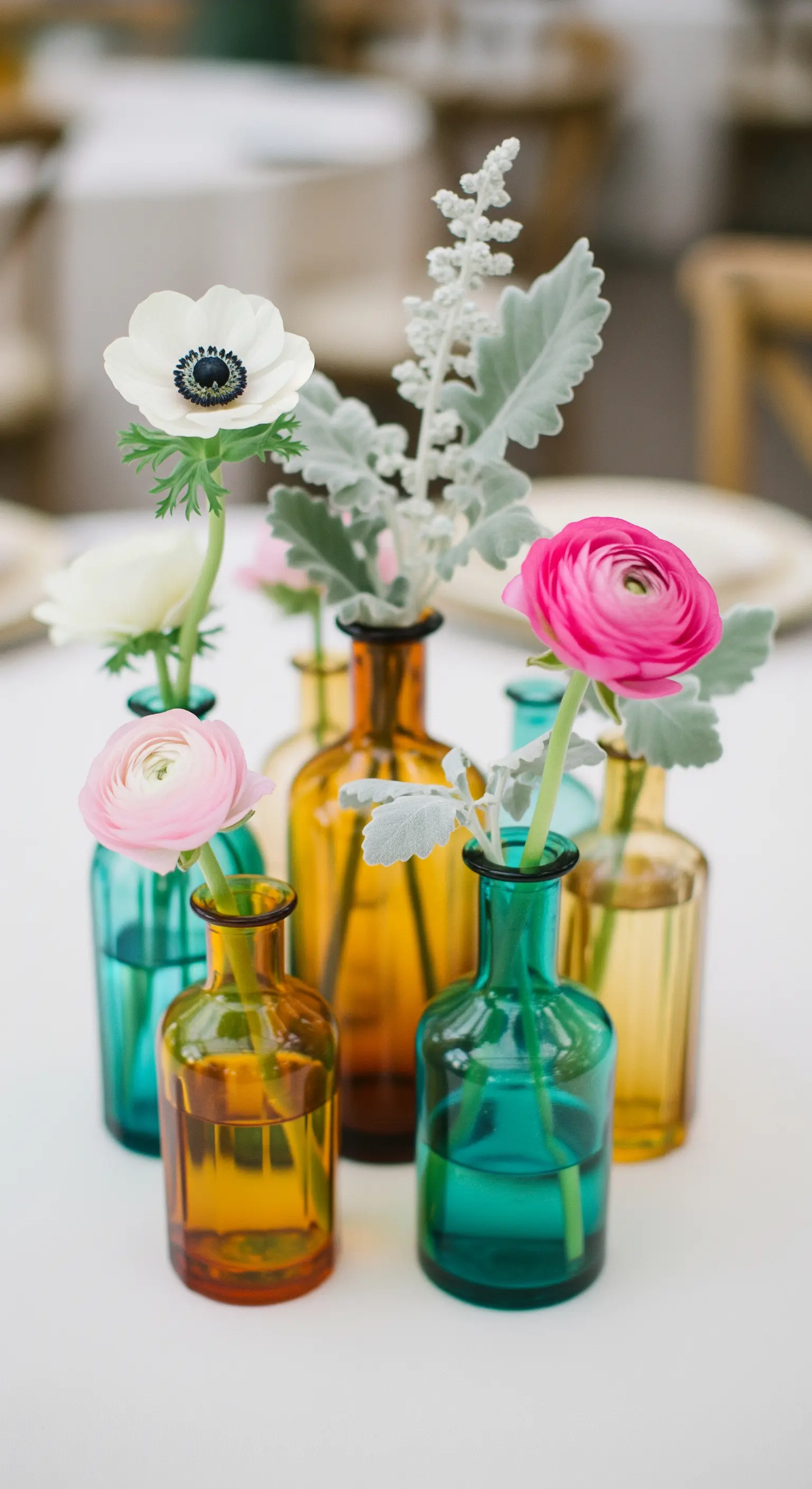 Fiori a stelo singolo, come ranuncoli, inseriti in piccole bottiglie di vetro colorato.