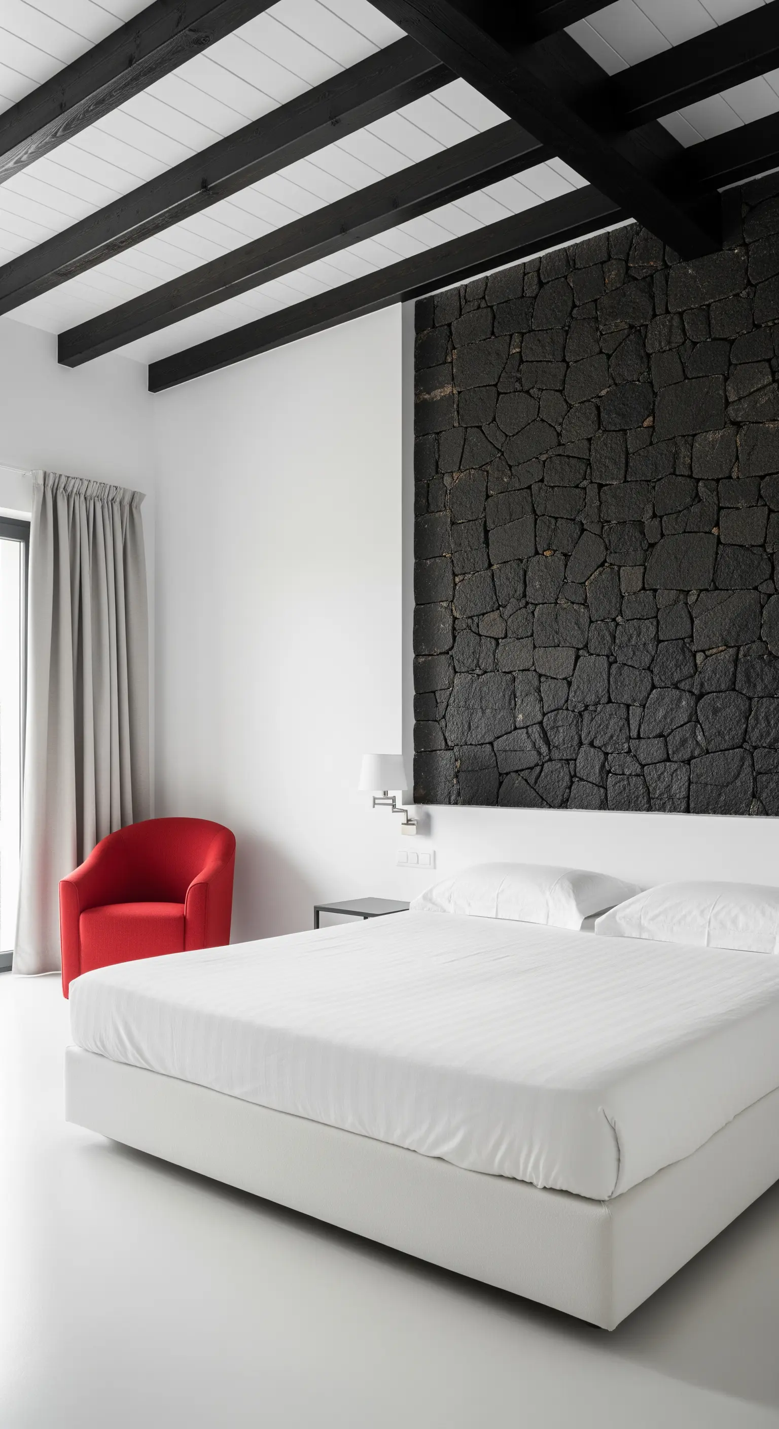 Camera da letto moderna con travi nere, pareti bianche, muro in pietra lavica e poltrona rossa.
