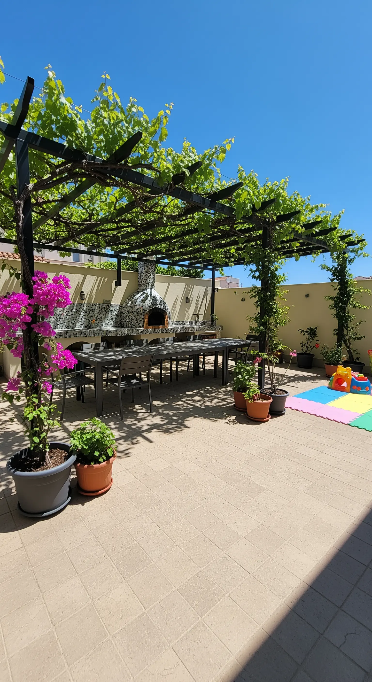 Patio con grande tavolo da pranzo, forno per pizza a mosaico e pergola di vite.