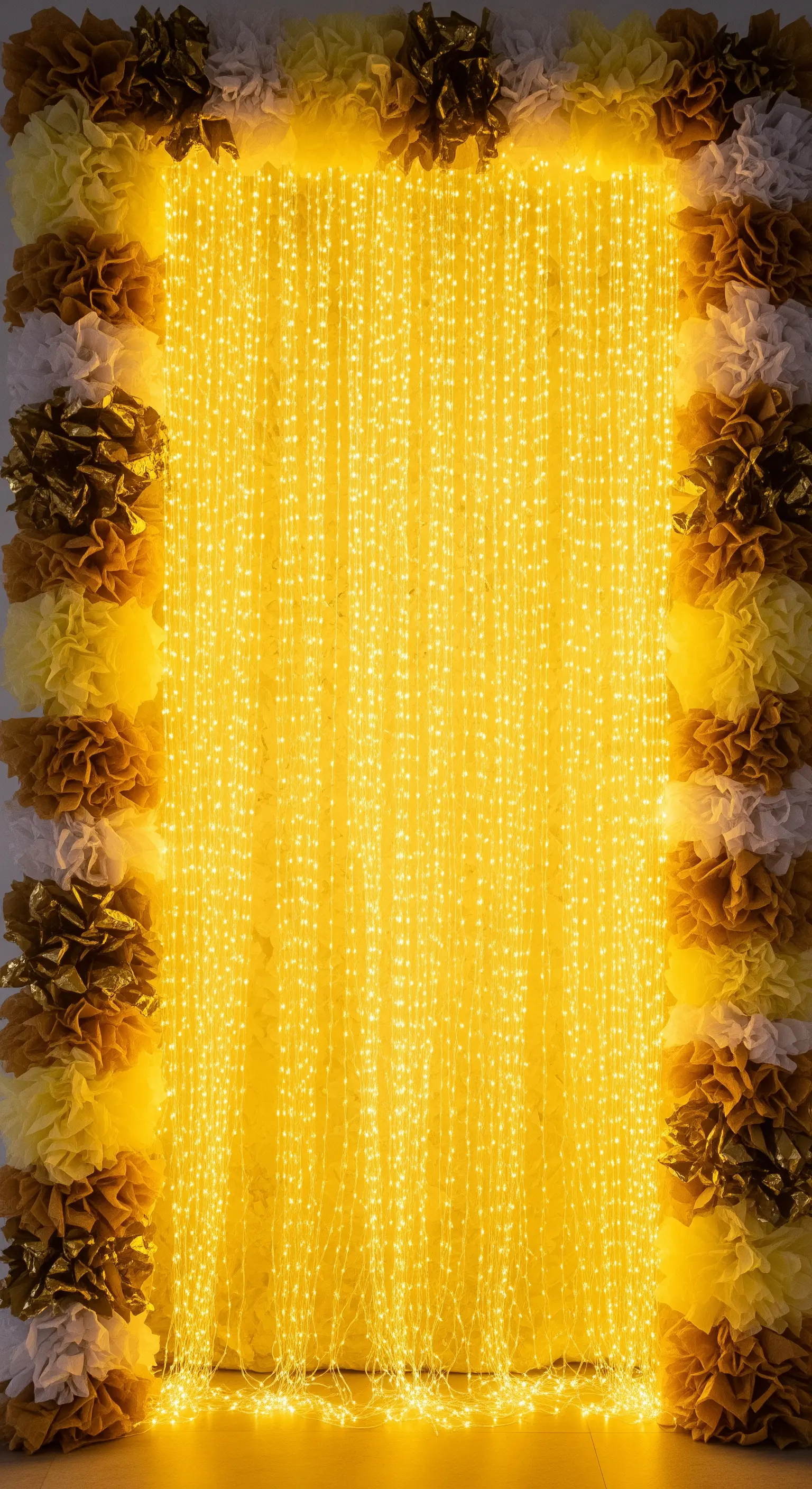 Cascata di lucine LED incorniciata da pompon di carta gialli, dorati e bianchi.