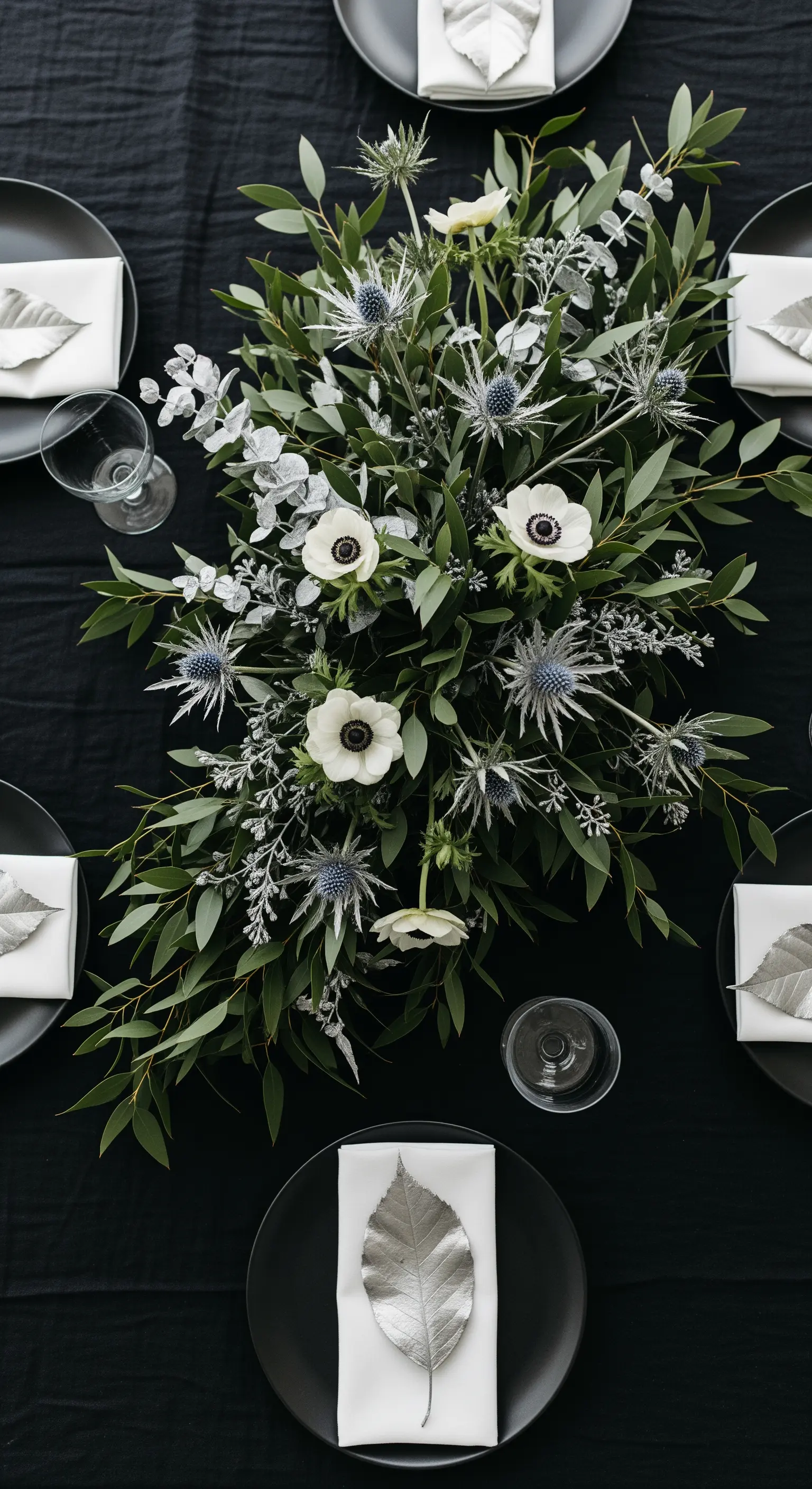 Centrotavola botanico con eucalipto, anemoni e dettagli argentati su tovaglia nera.