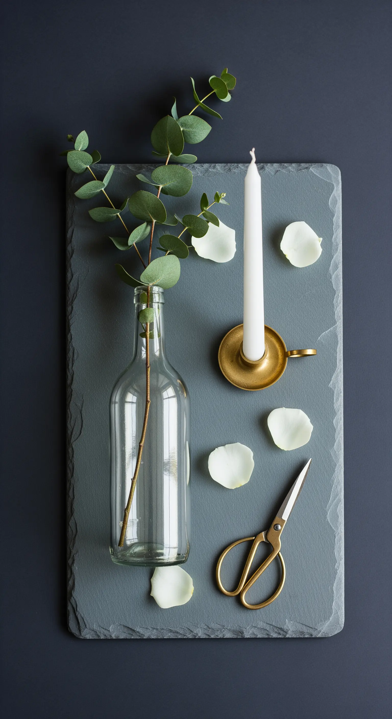 Composizione minimalista con bottiglia, ramo di eucalipto e candelabro dorato su ardesia.